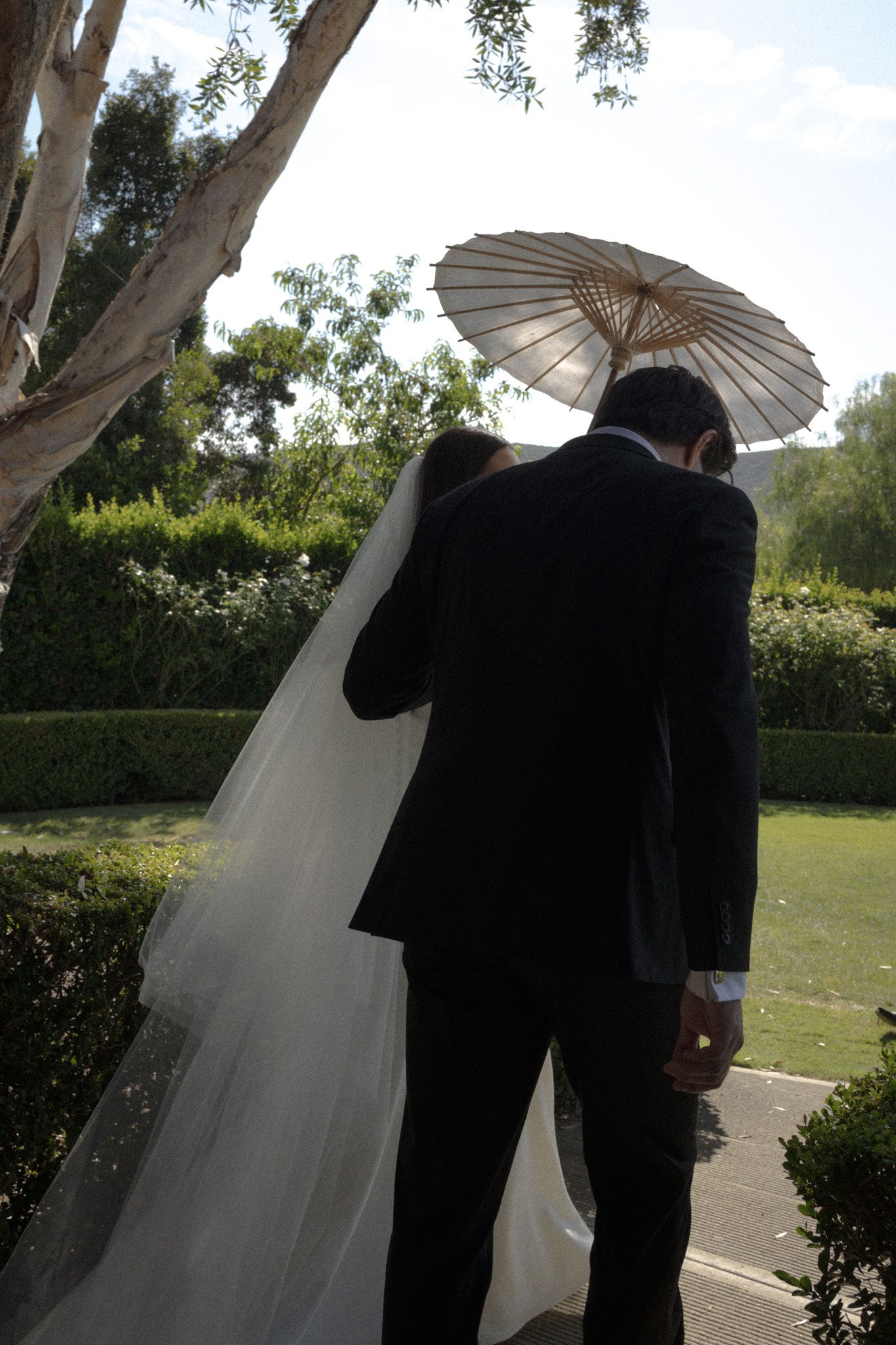 Wedding couple walks under white parasols in an outdoor garden setting.