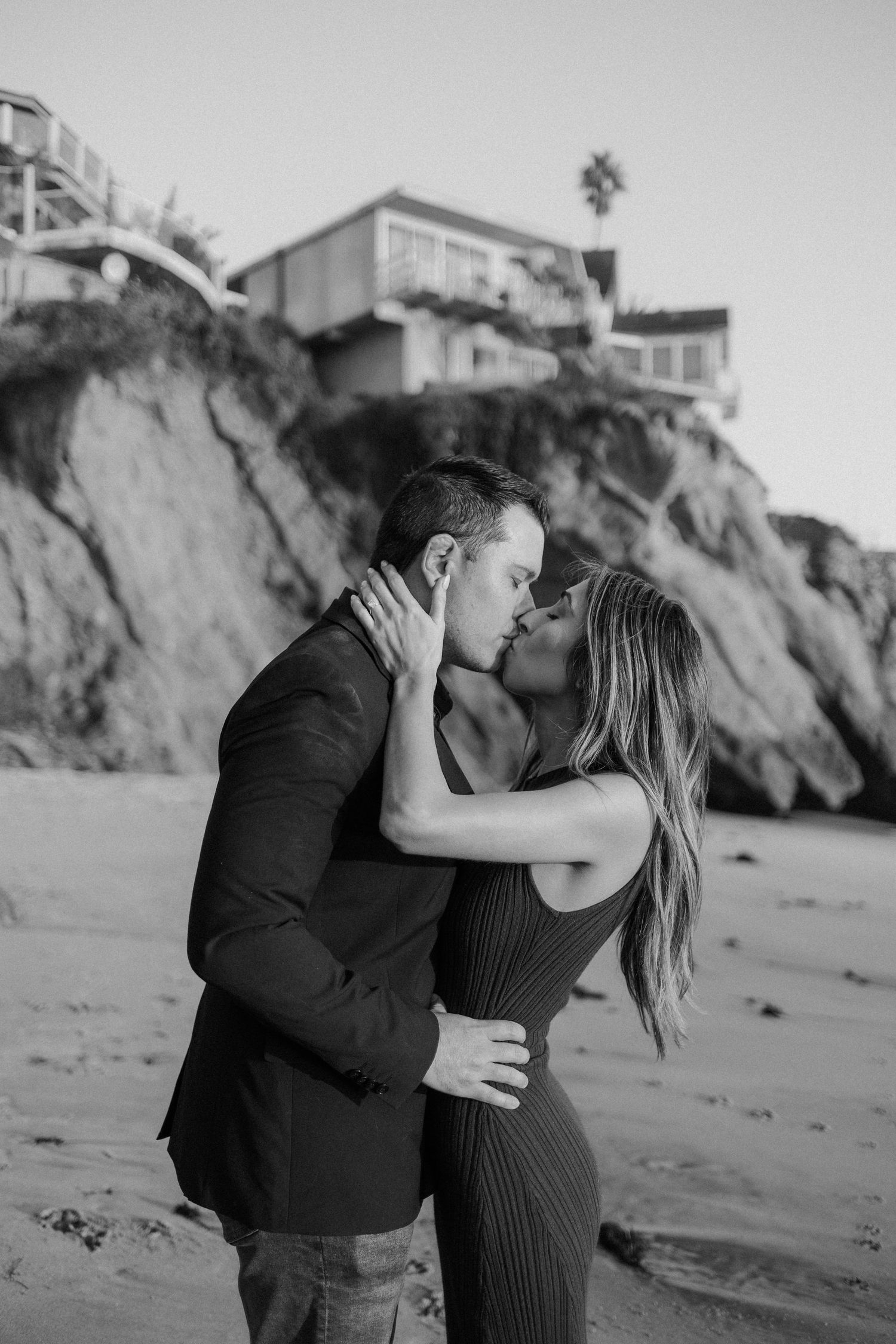 A romantic black and white photo of a couple sharing an intimate moment on a beach with cliffside homes in background.