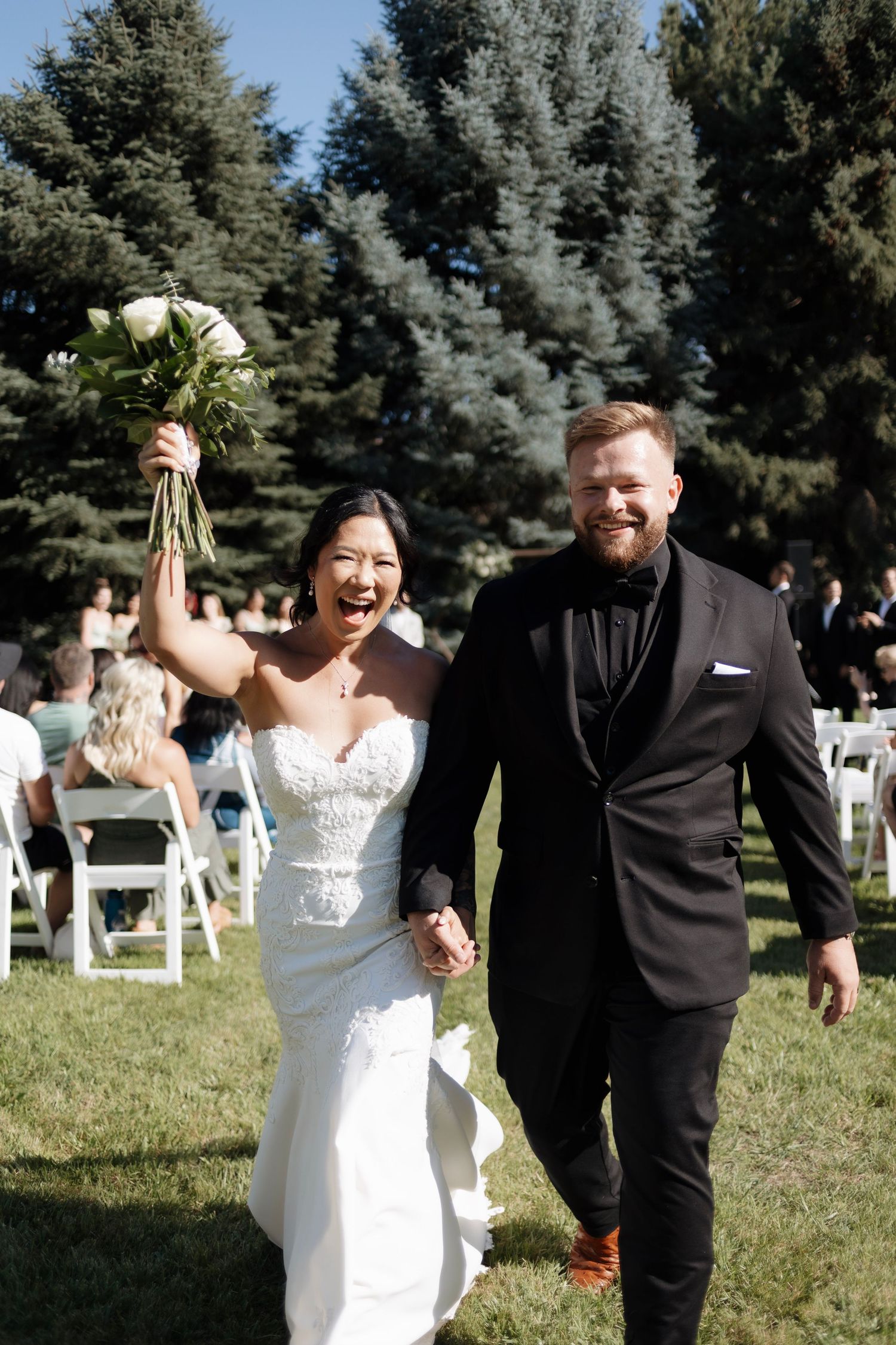 A bride and groom share joyful moments walking together on a grassy lawn with white chairs and evergreen trees in background.