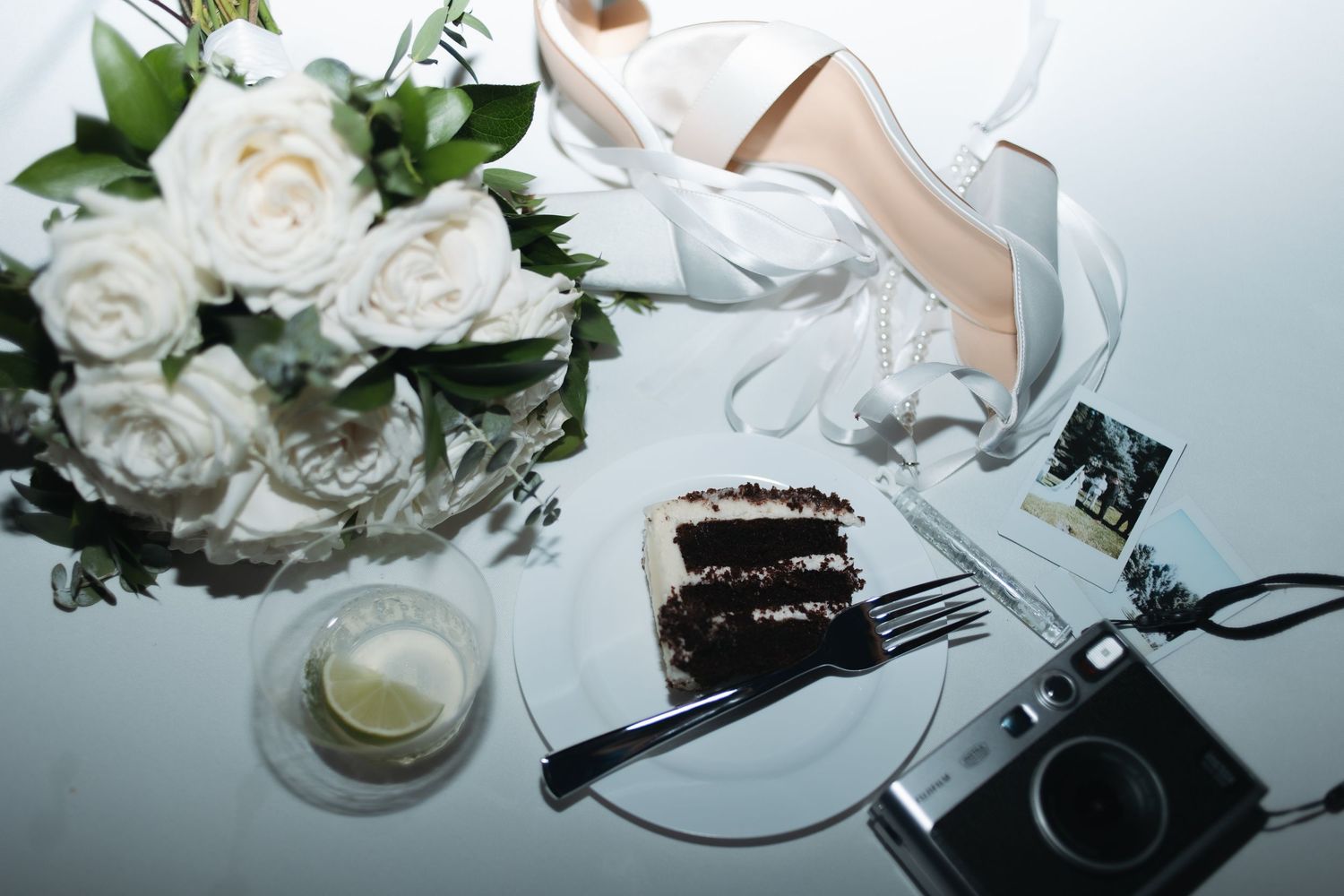 Wedding scene with white roses, camera, and slice of chocolate cake on reflective surface.