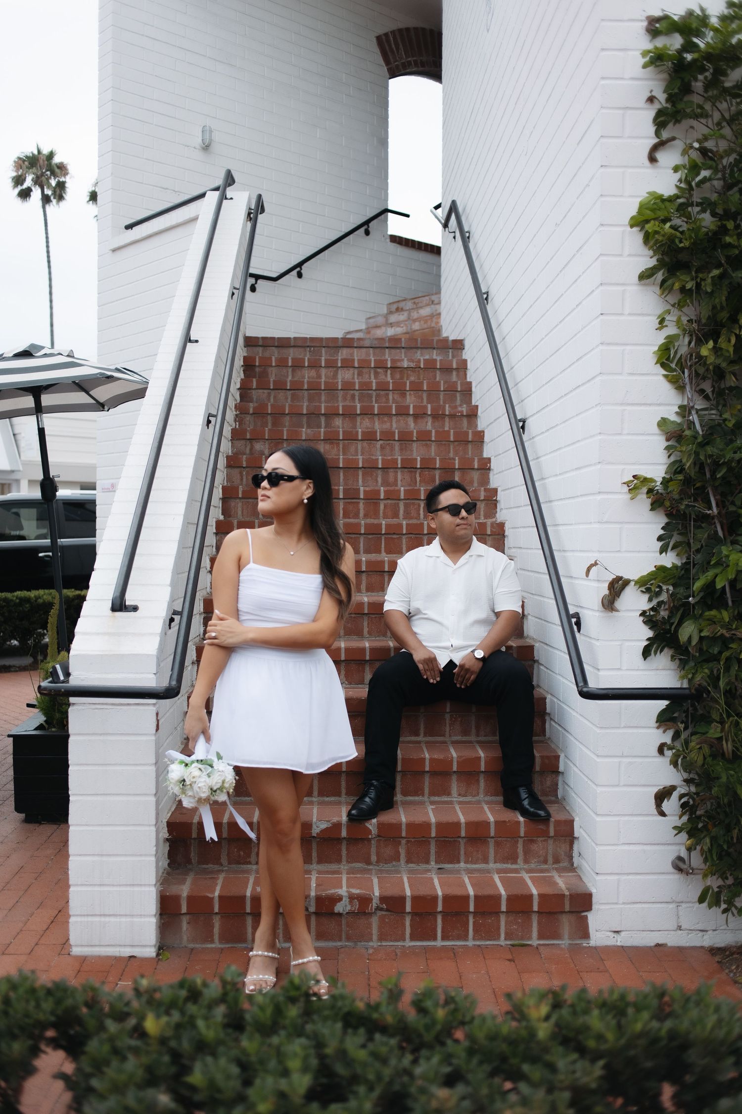 Couple relaxing on brick steps against white wall in casual attire.