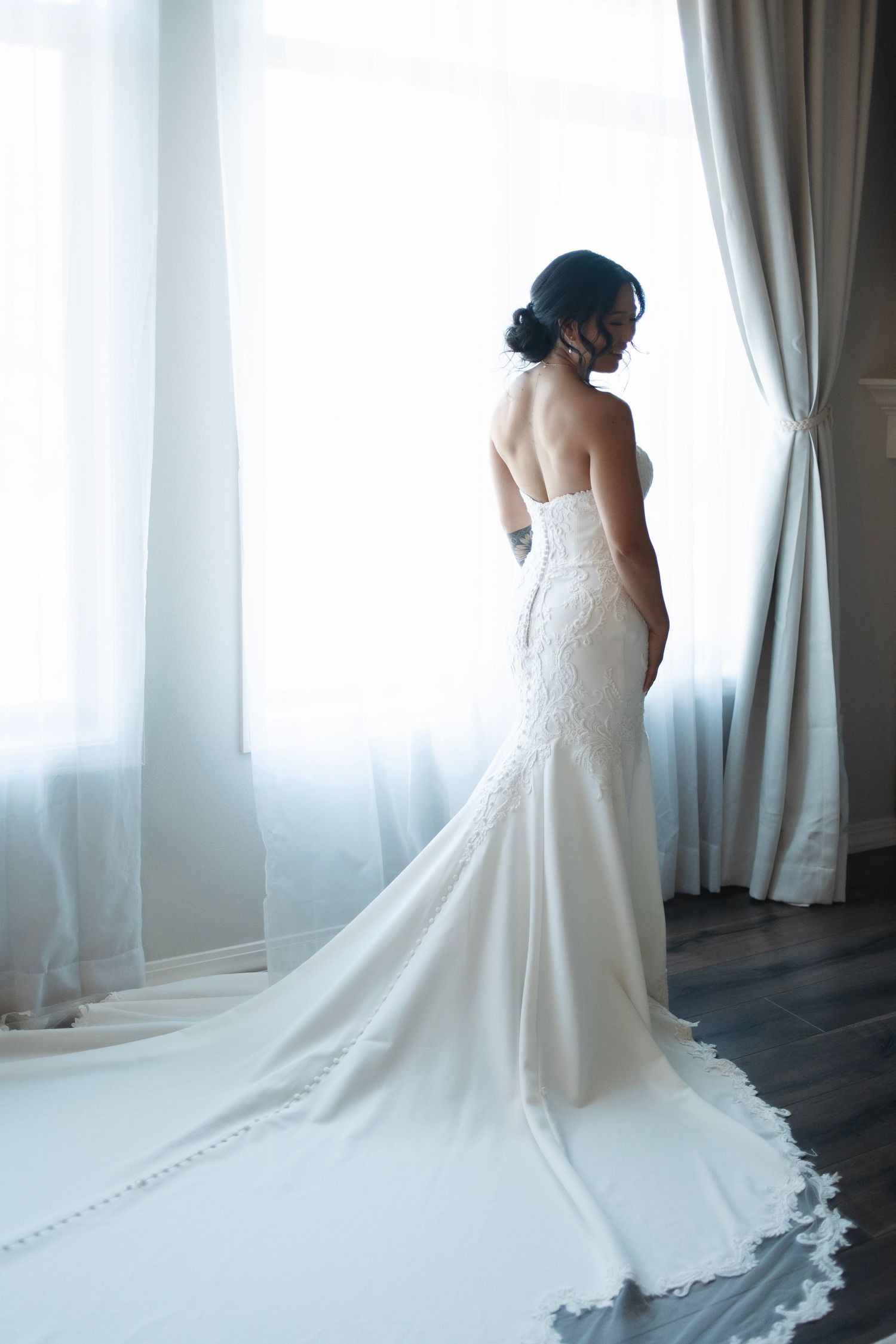A bride in an elegant strapless mermaid wedding gown stands by a window, showing the dress's flowing train and lace details.