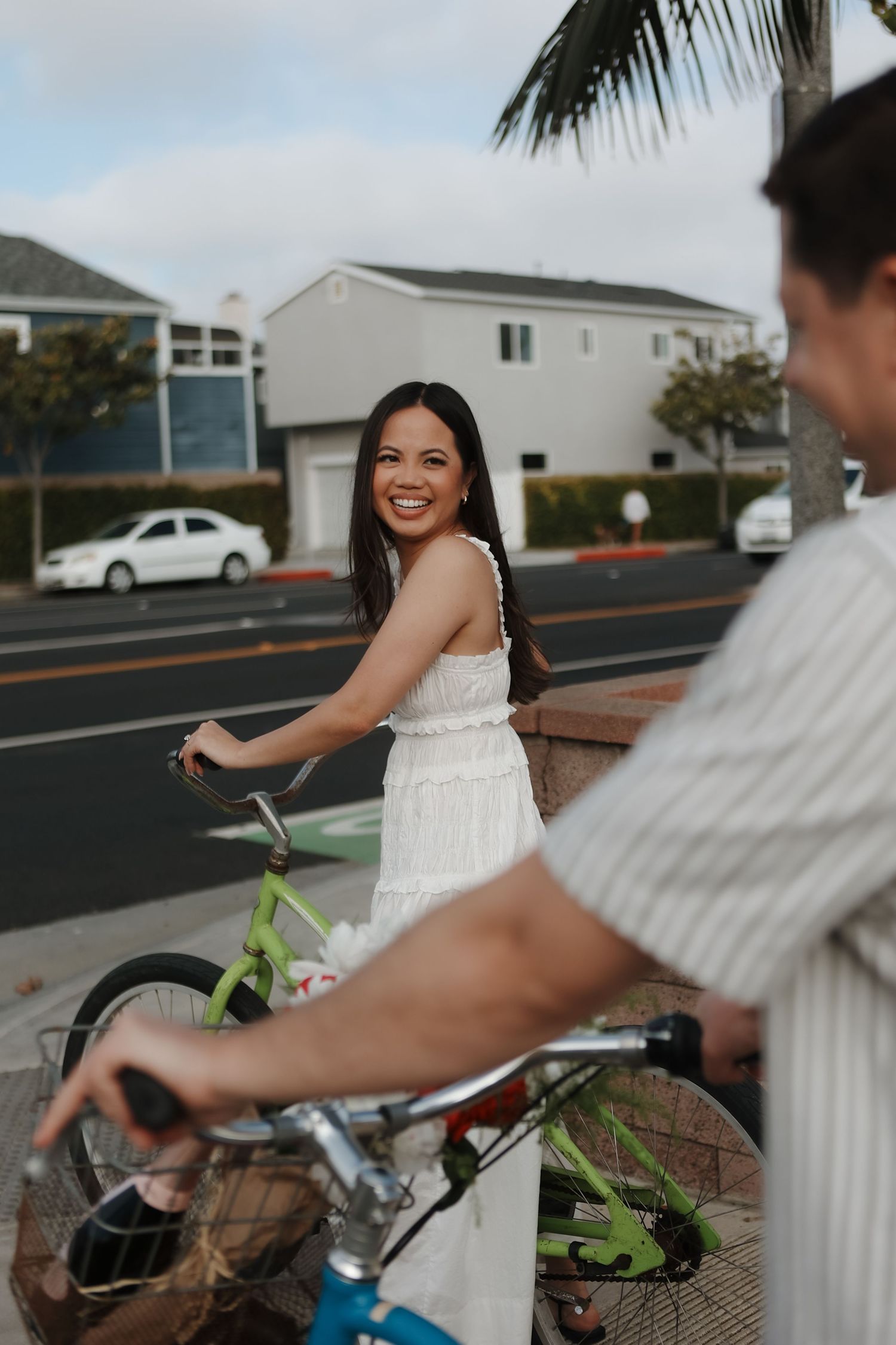 A couple rides bicycles together on a residential street with palm trees and houses in the background.