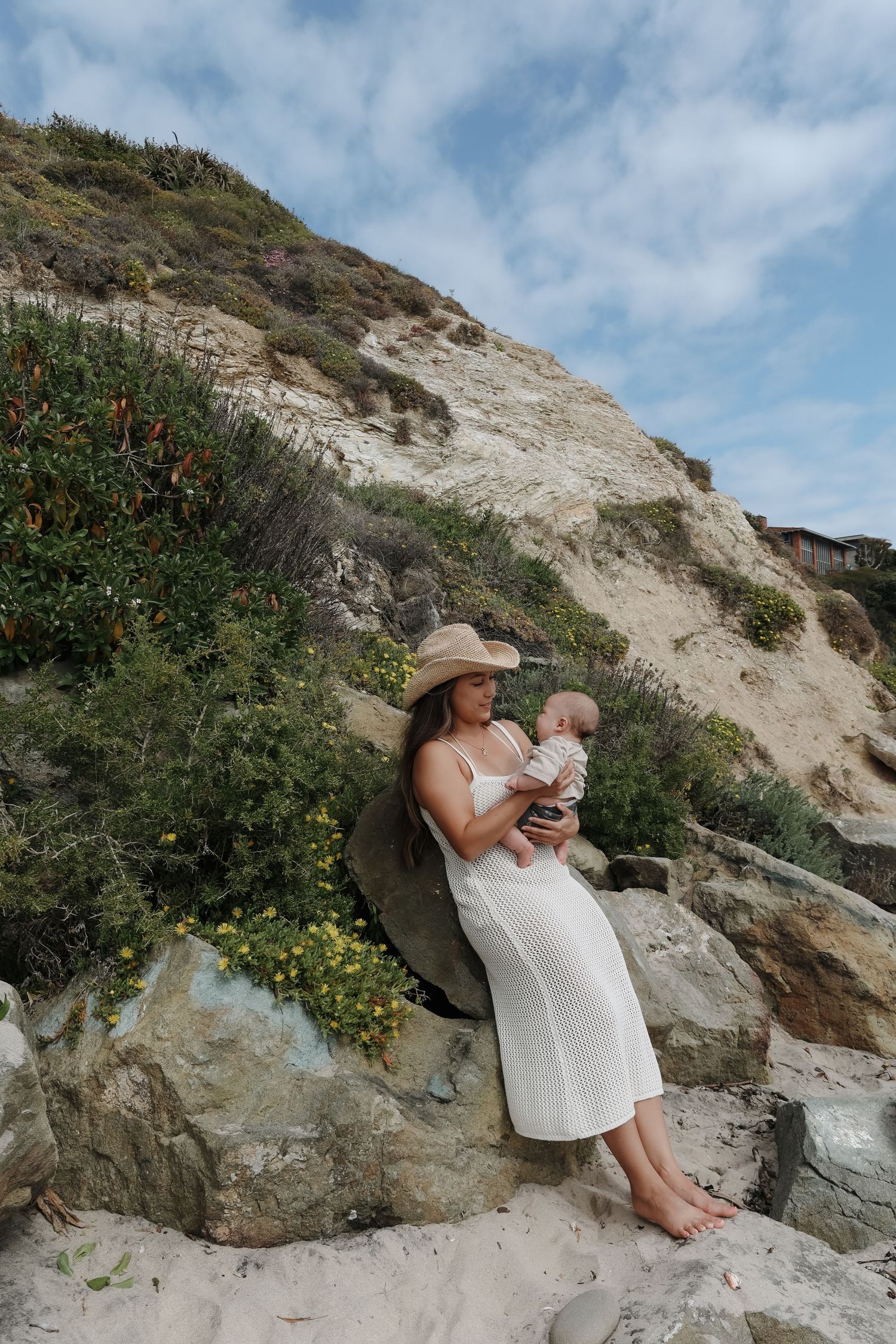 A person in a sun hat and white dress sits on coastal rocks with cliffs and blue sky in the background.
