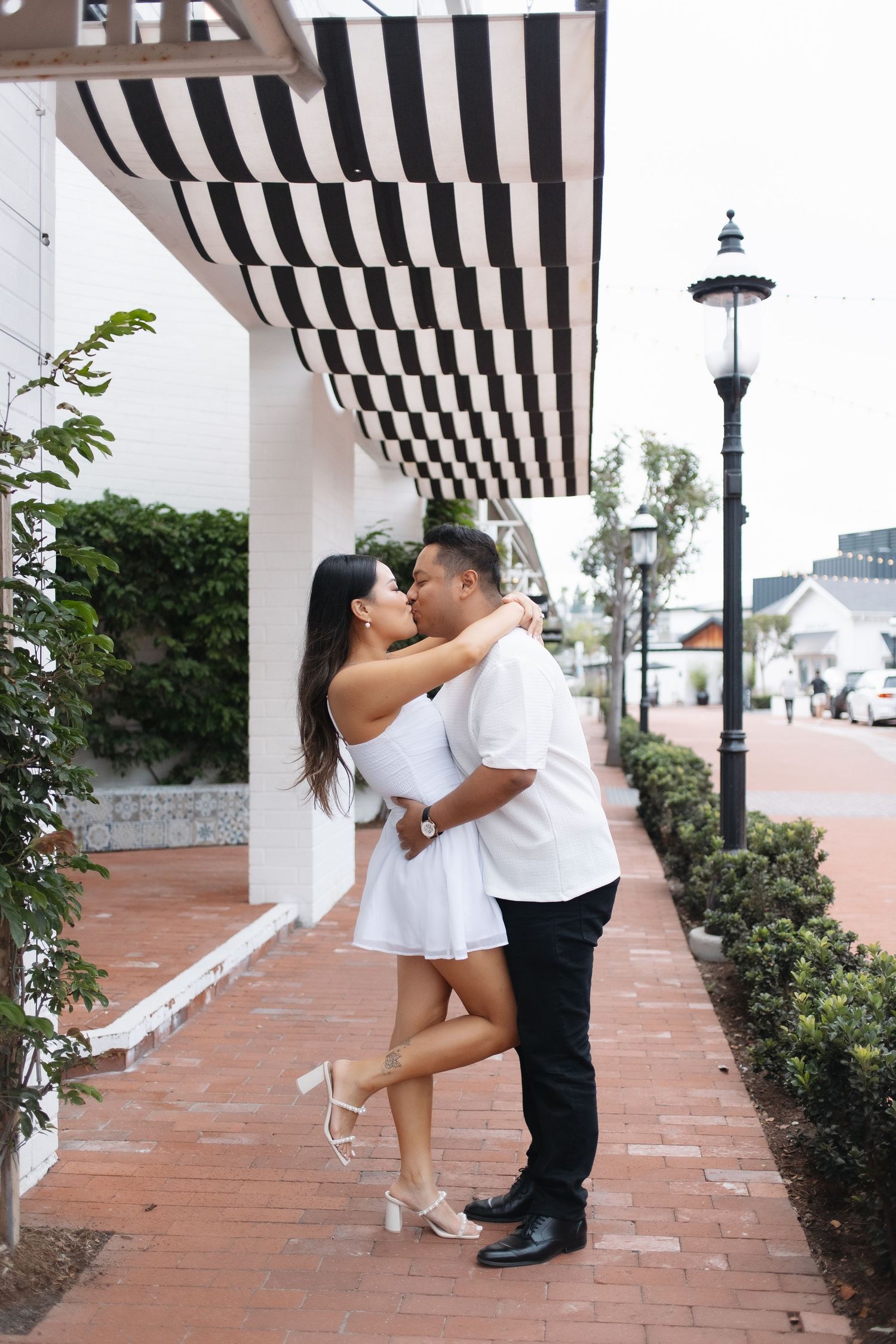 A couple shares a romantic dip-style kiss under a black and white striped awning on a brick patio.