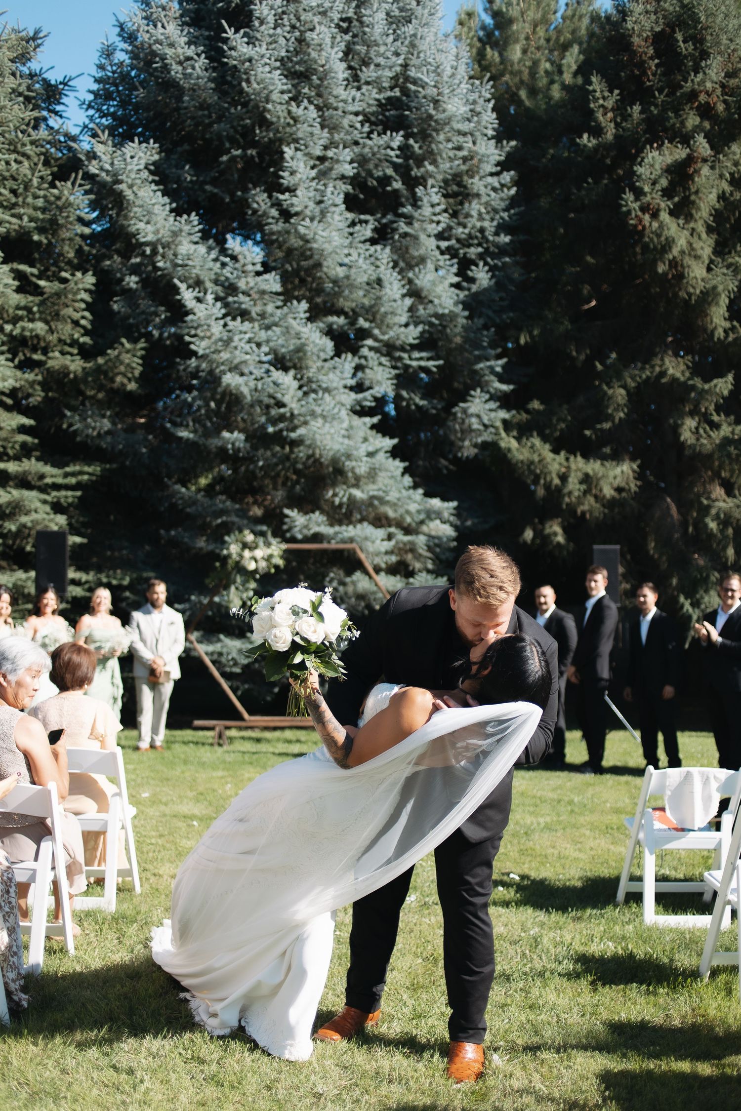 Outdoor wedding ceremony setup with white chairs on grass surrounded by evergreen trees in natural lighting.