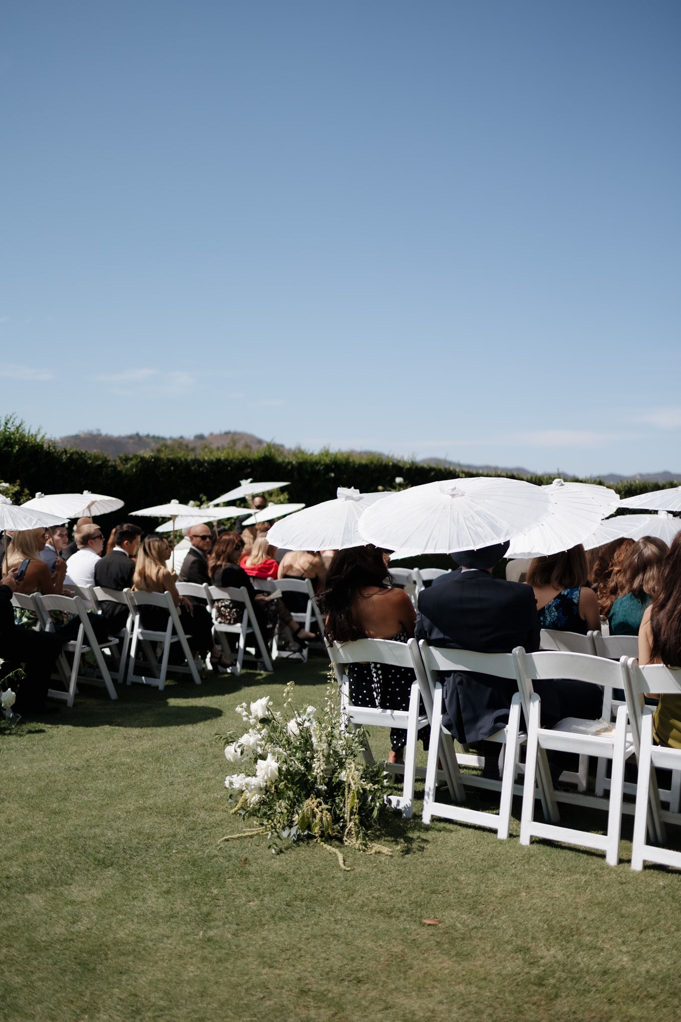 Wedding guests seated in white chairs with parasols at an outdoor ceremony under blue skies.