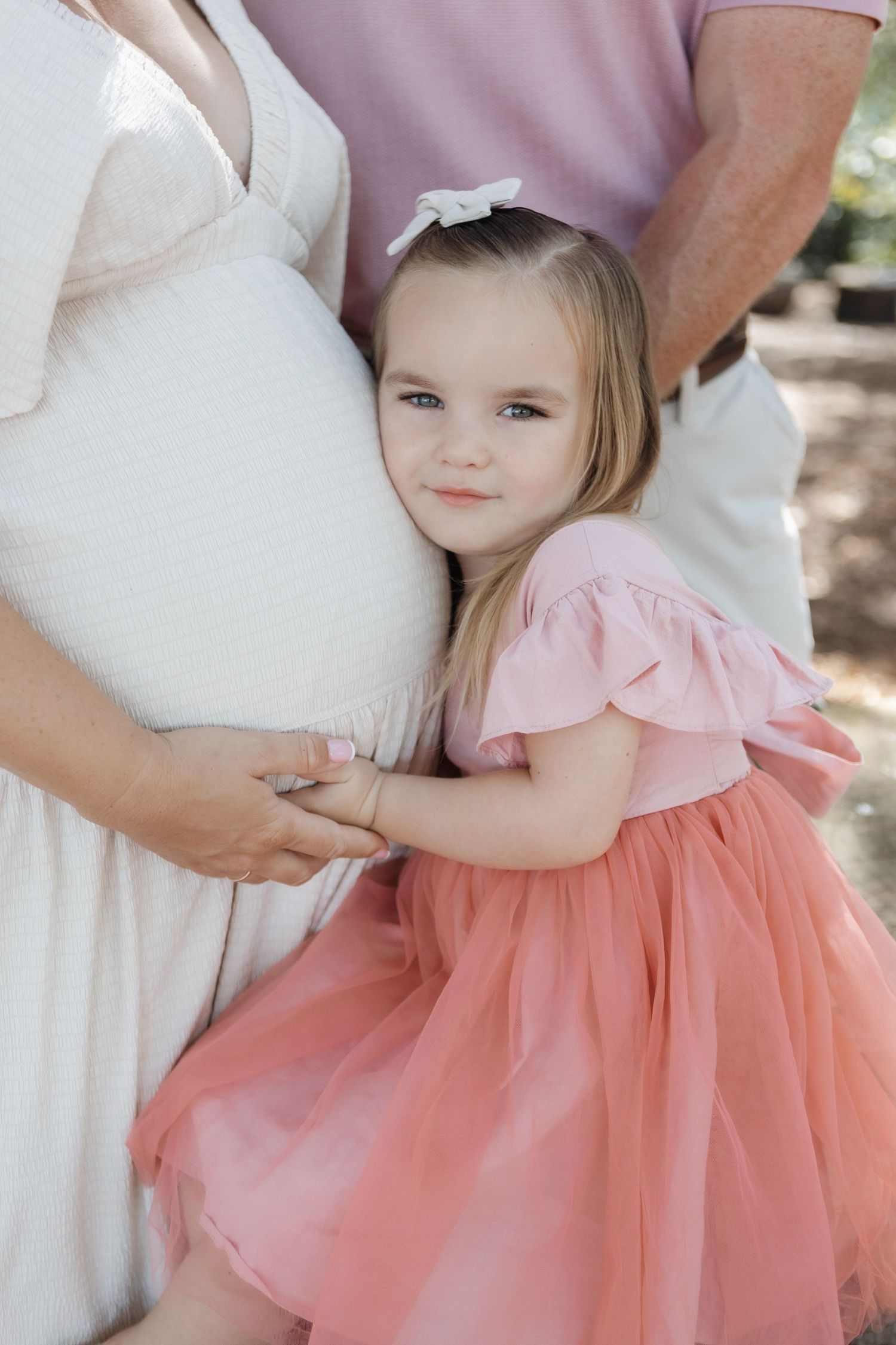 A child wearing a pink tulle dress stands close to someone in a white dress during an outdoor photo session.