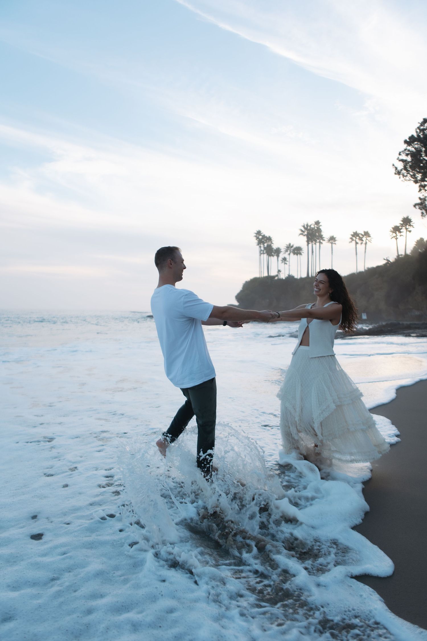 Couple dancing and splashing in ocean waves on a scenic beach with palm trees and cliffs at sunset.