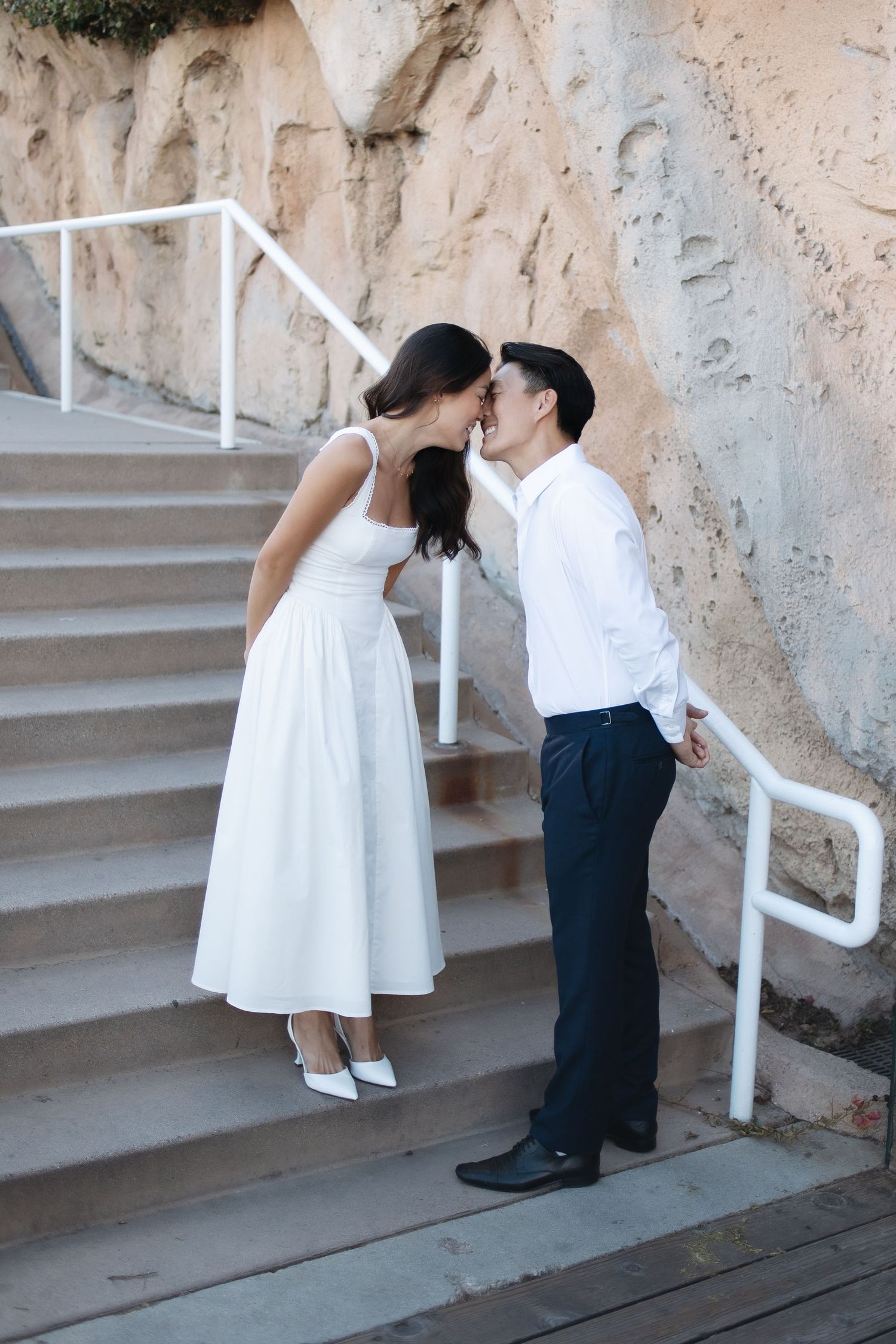 Two young children in white and dark clothing embrace and kiss sweetly on stone stairs with a white railing.