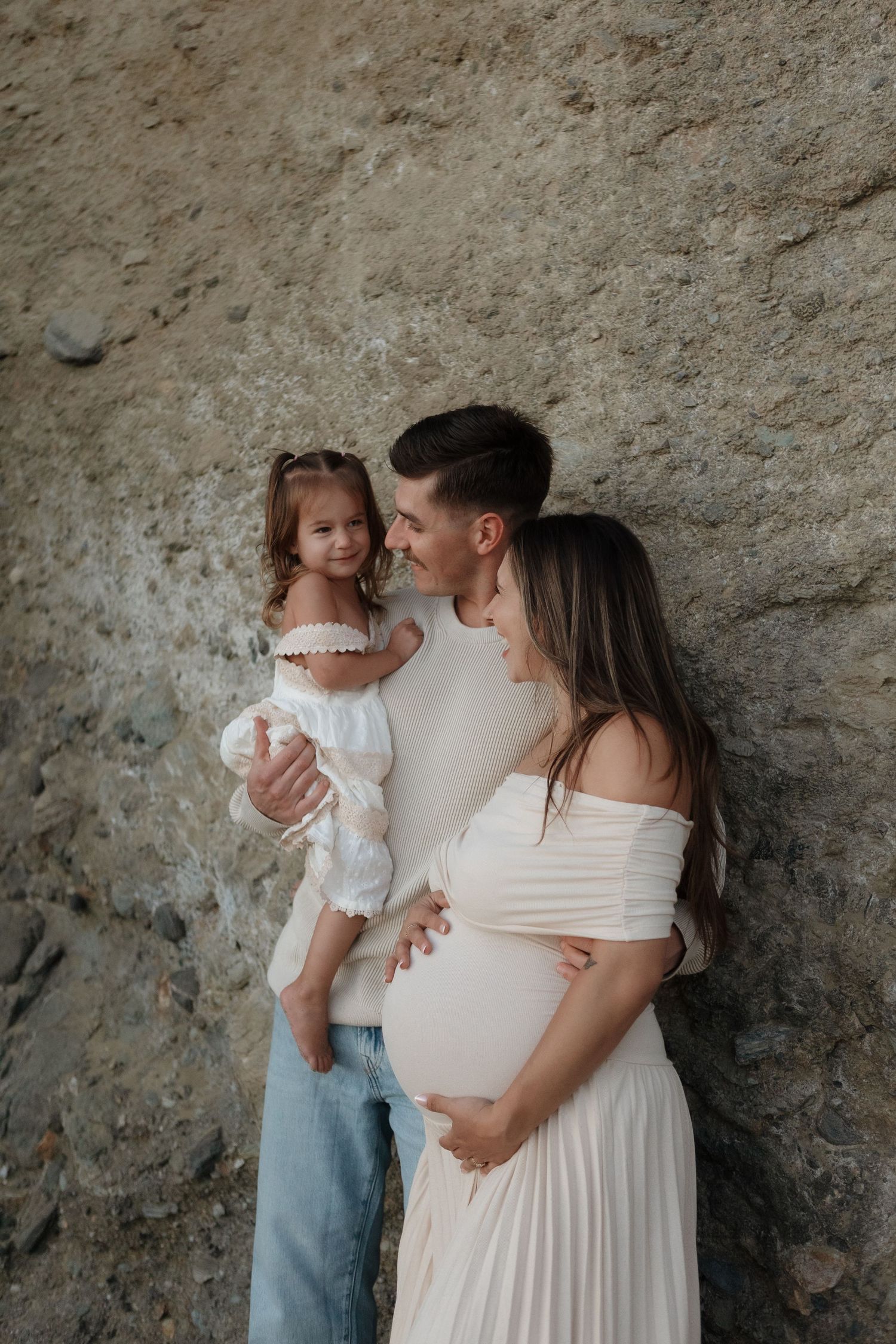 A family in white outfits shares tender moments against a textured stone wall backdrop.