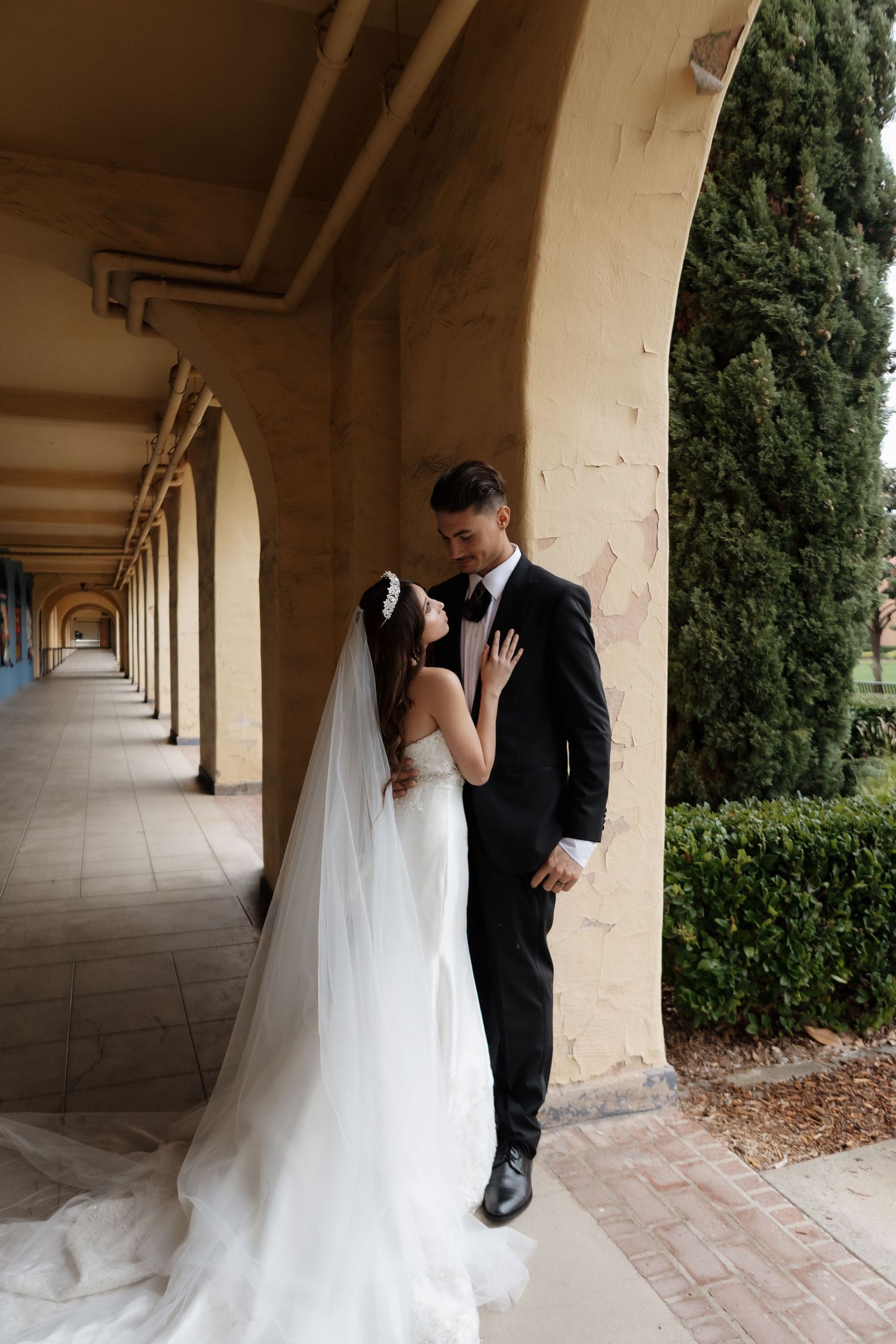 A romantic wedding couple shares intimate moments under a beautiful columned walkway with cypress trees in the background.