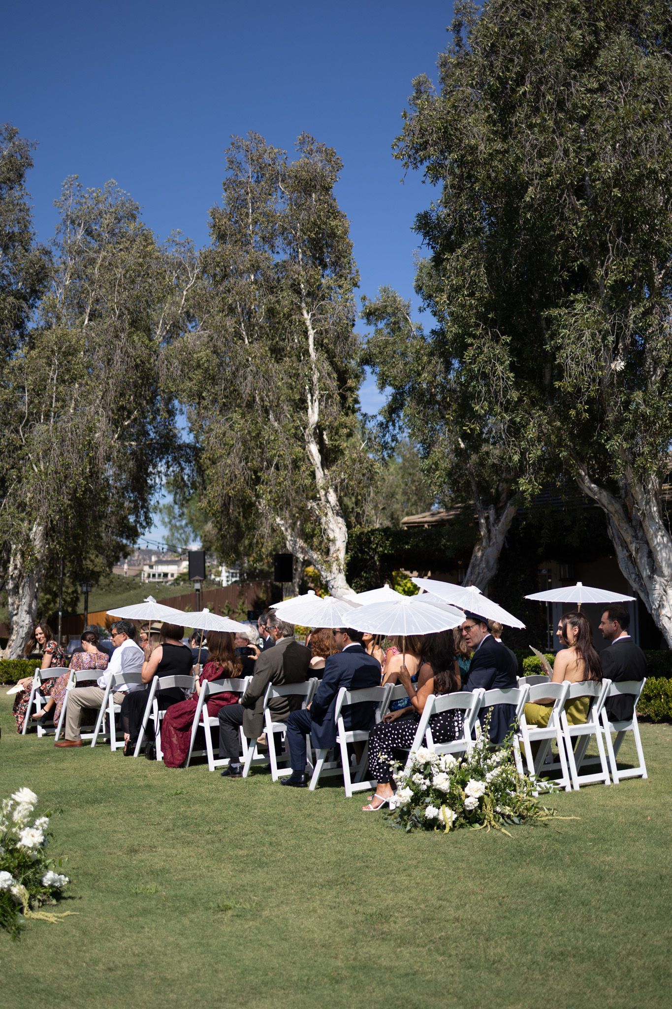 Outdoor wedding ceremony setup with white chairs arranged on a grassy lawn under tall eucalyptus trees on a sunny day.