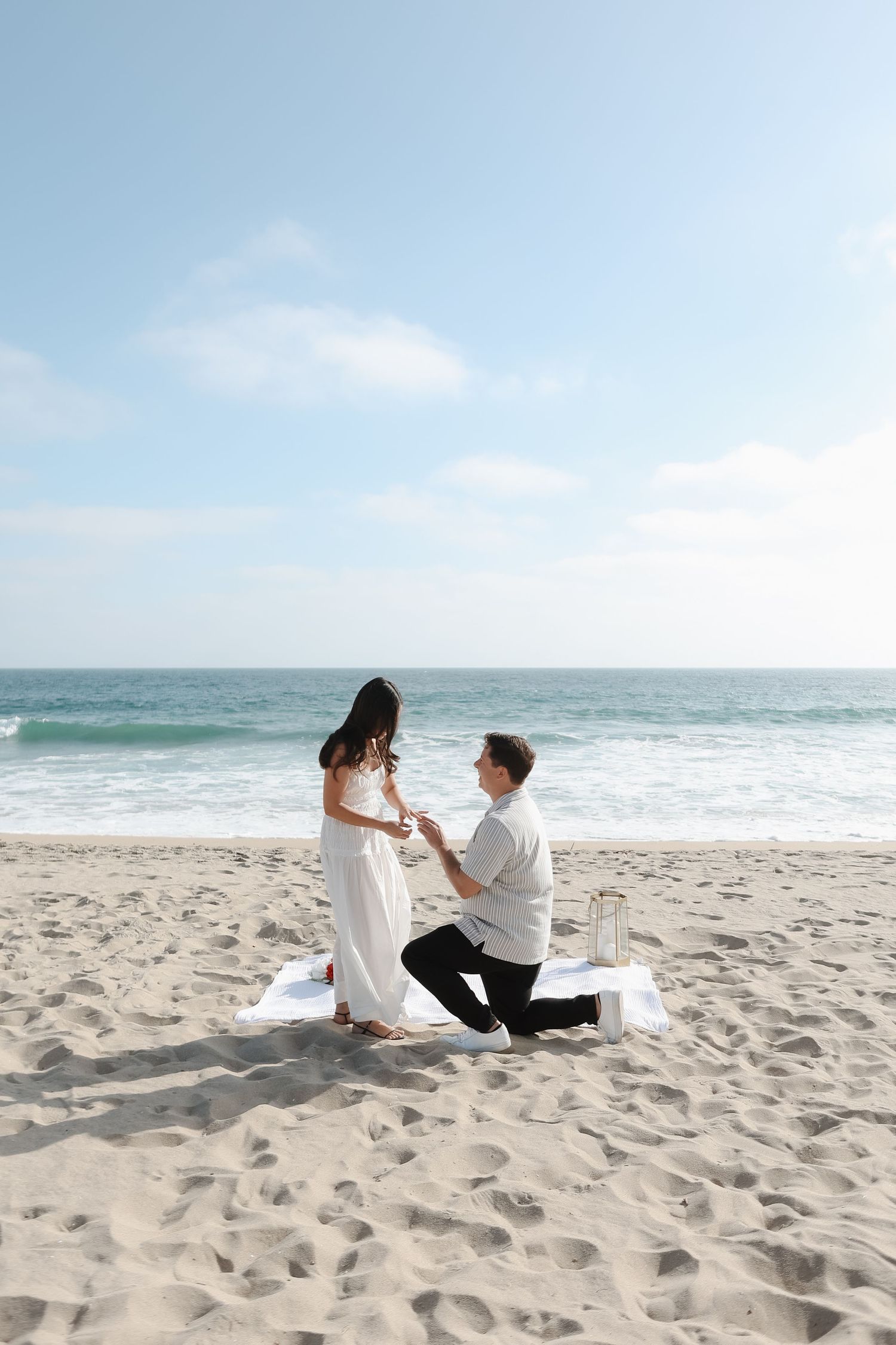 A couple sits together on a sandy beach with ocean waves and blue sky in the background during a romantic moment.