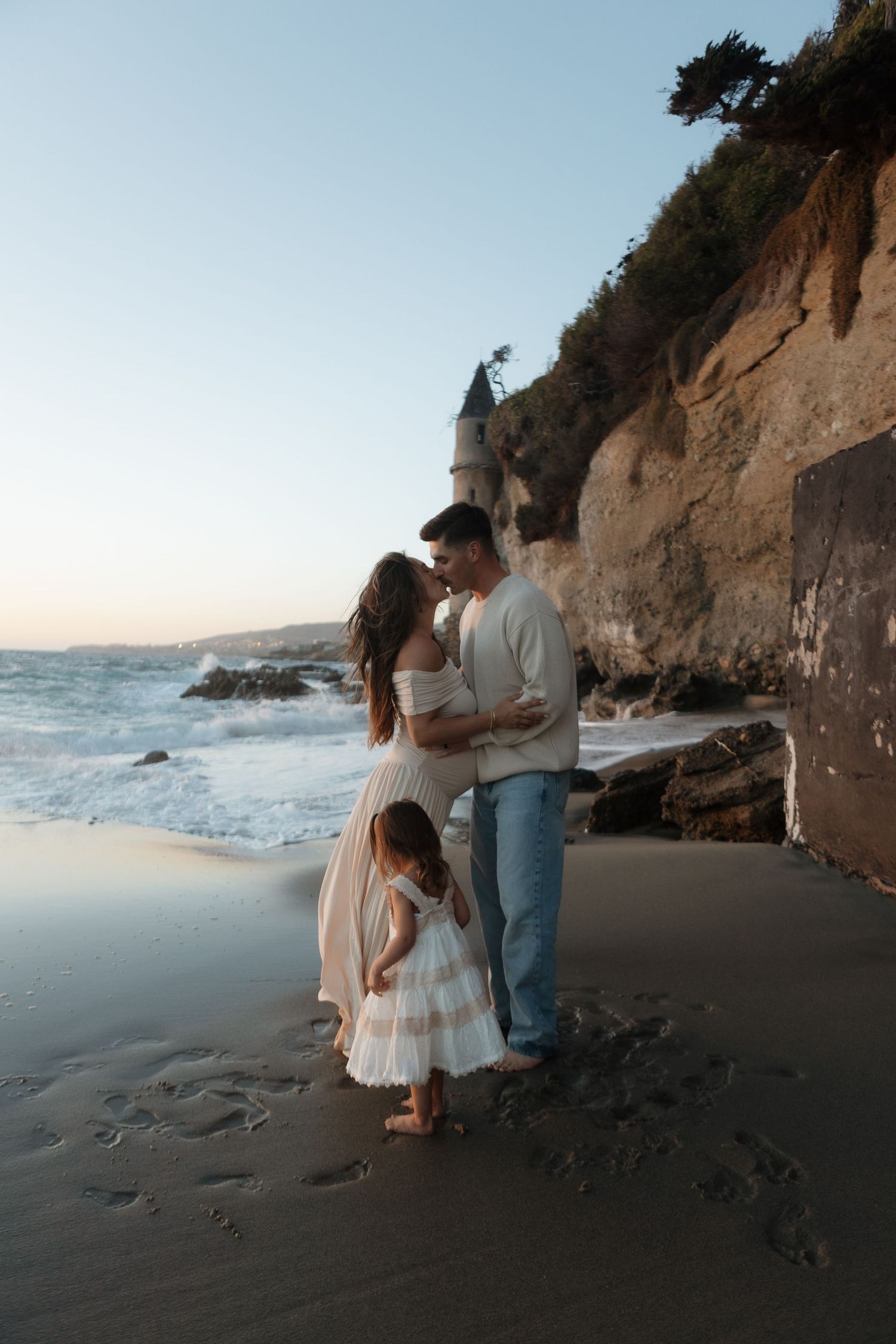 A couple shares an intimate moment on a scenic beach at sunset with dramatic cliffs in the background.