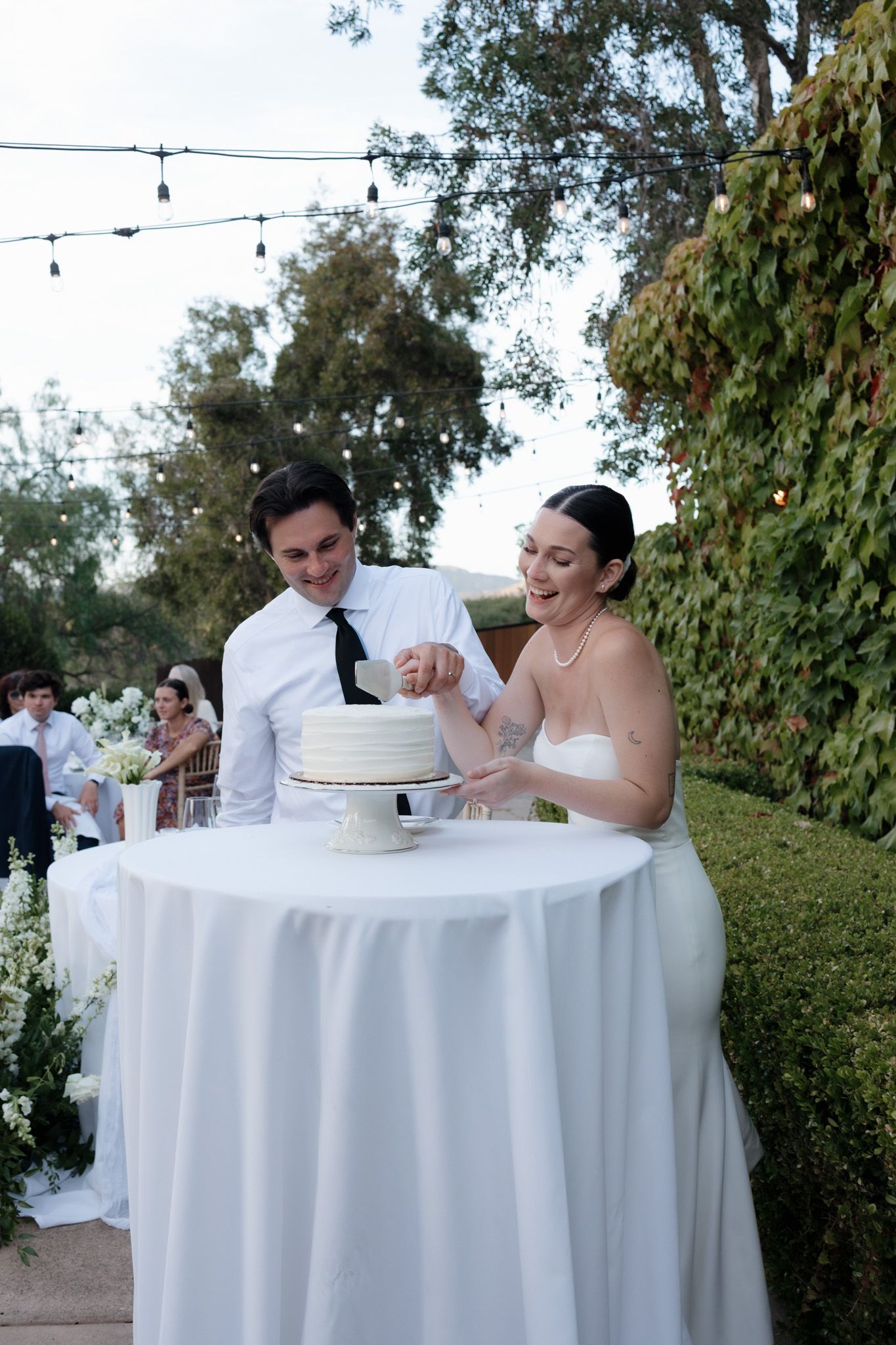 A couple in wedding attire shares a playful moment while sitting at a round table with white linens outdoors.