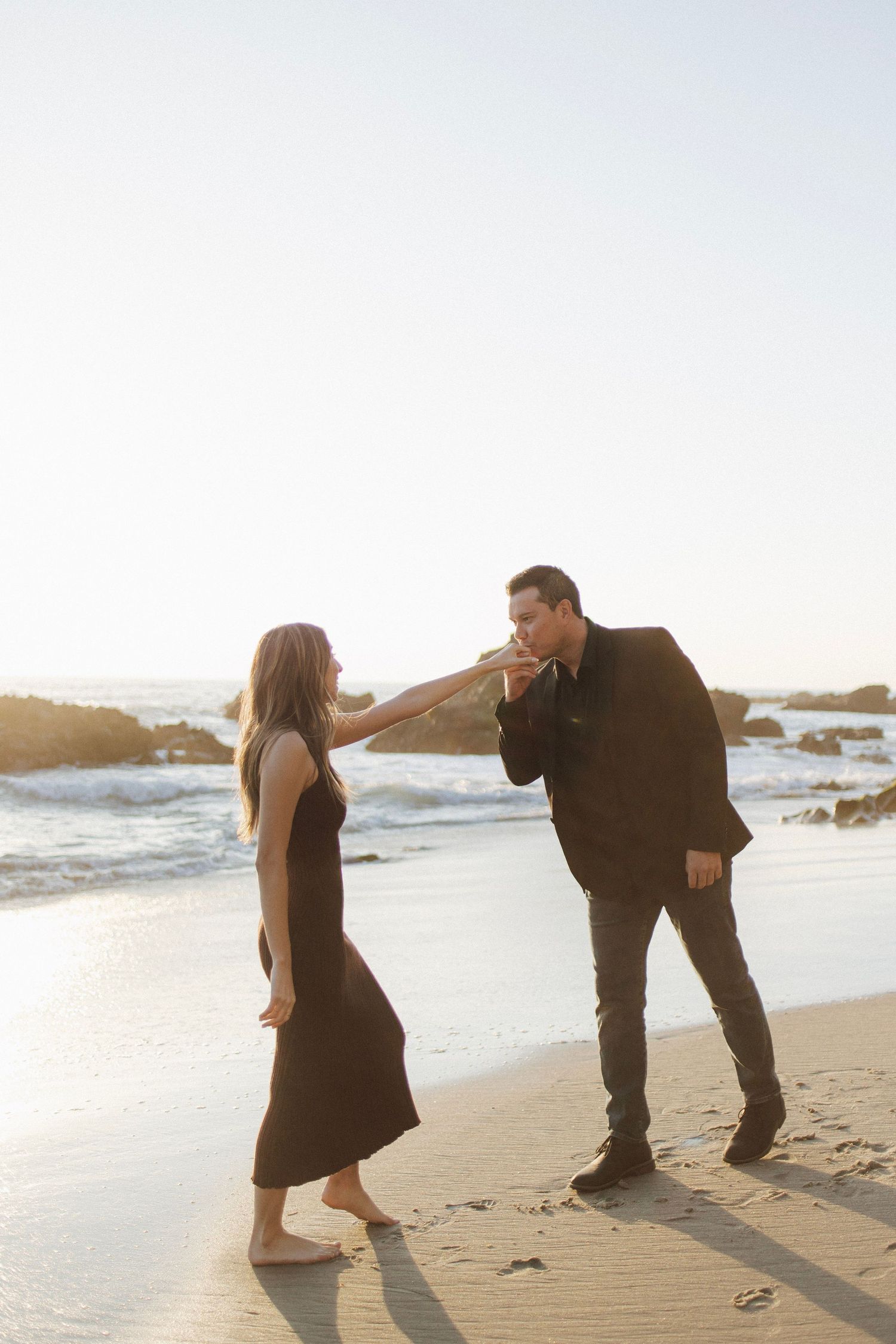 A couple shares a romantic moment on a beach at sunset as waves crash on the rocky shore in the background.