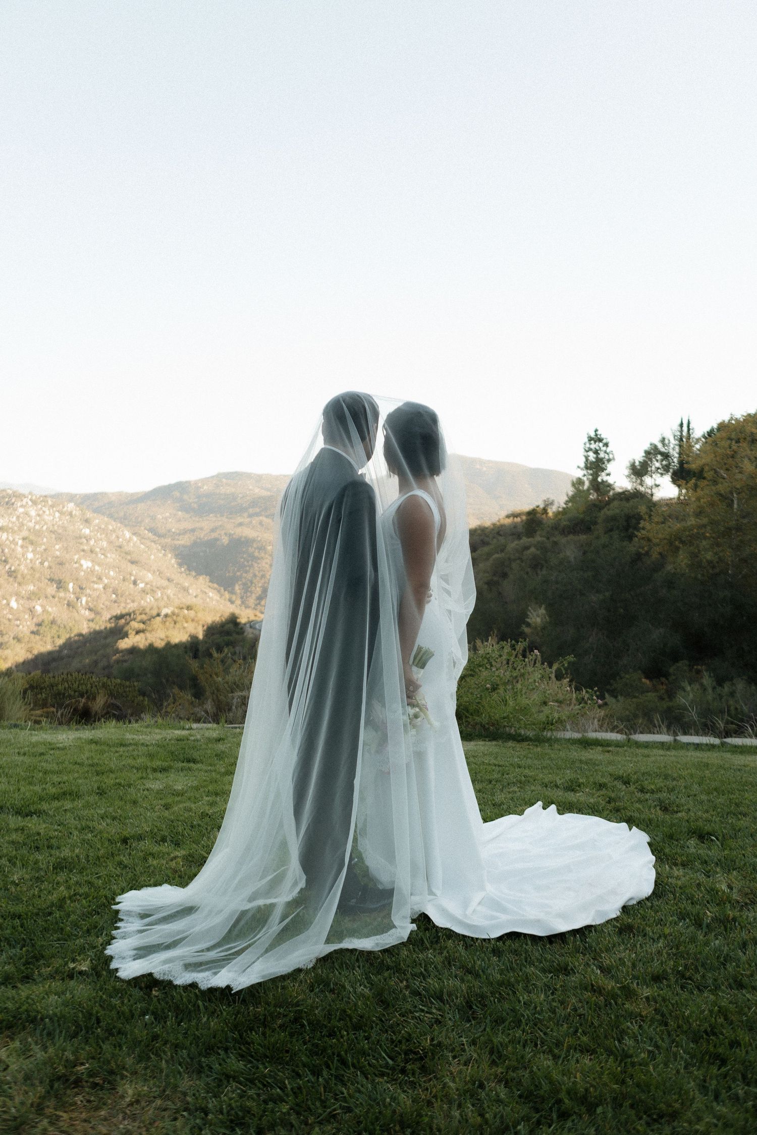 Two people in flowing white wedding dresses and veils stand together on a grassy hilltop with mountains in the background.
