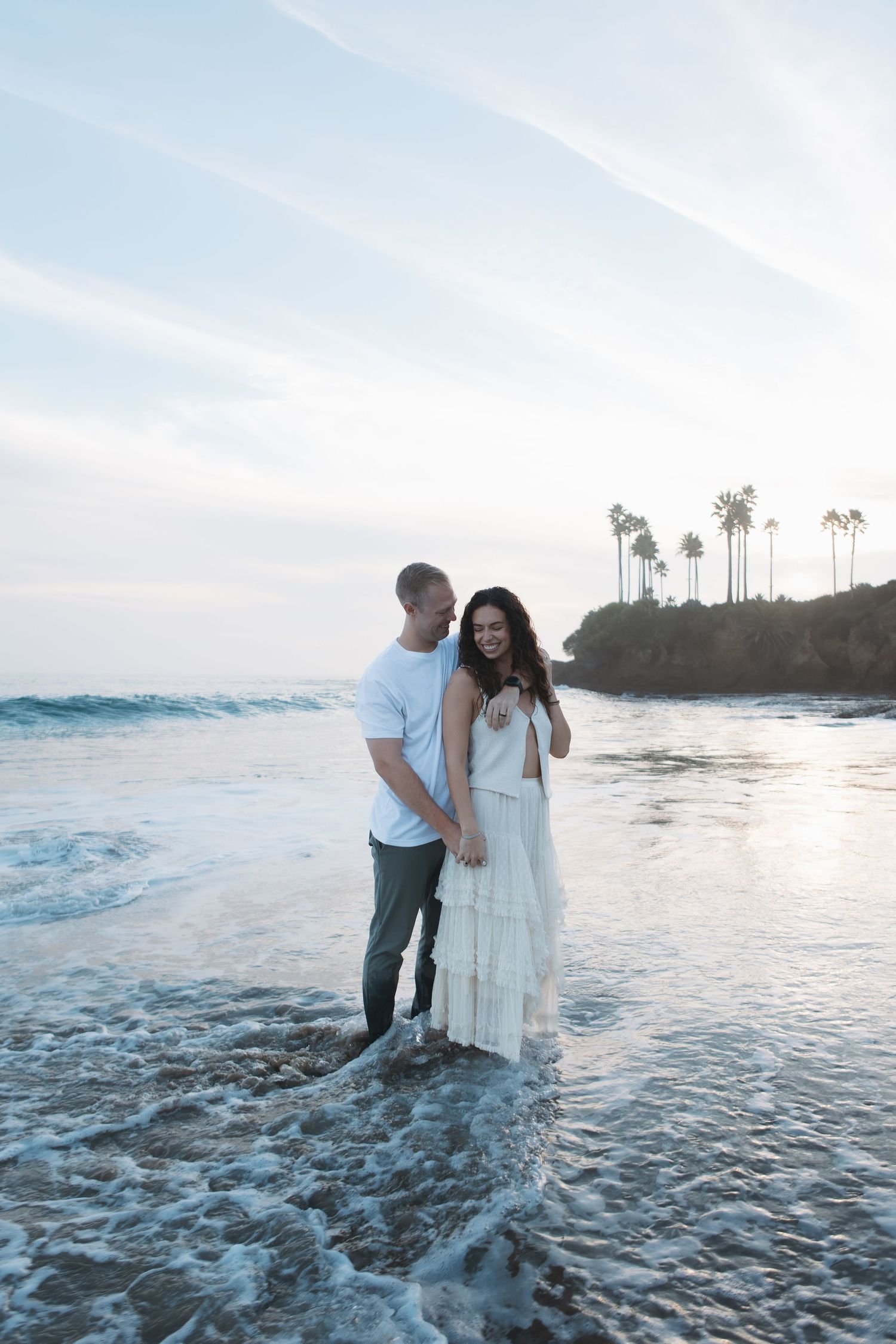 A couple stands together in the ocean waves at sunset with palm trees silhouetted against a blue sky in the background.