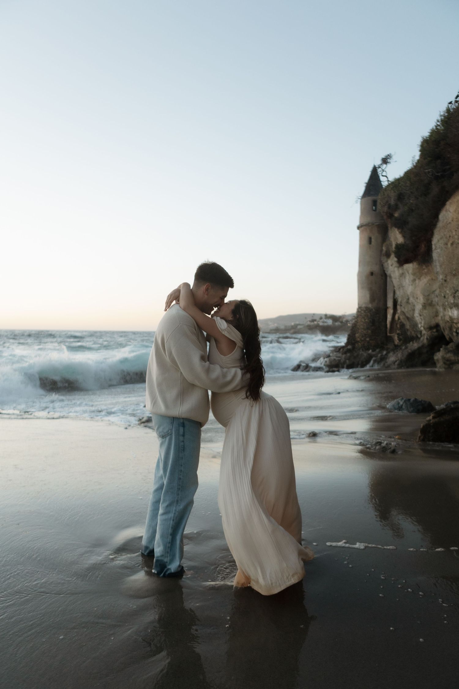 A romantic couple embraces on a misty beach with dramatic cliffs and crashing waves in the background.