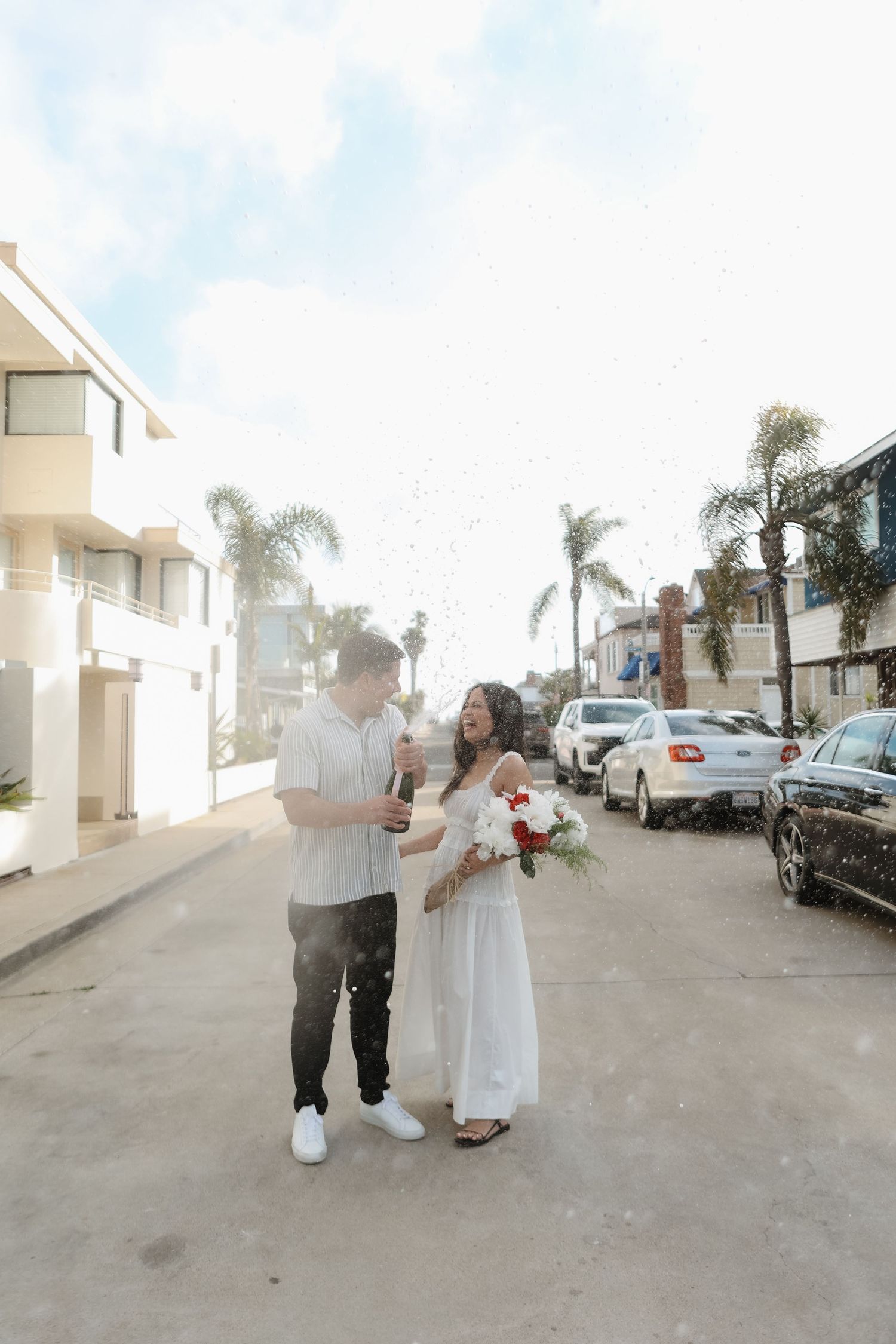 Romantic couple in white outfits dancing together on a sunny day with palm trees and buildings in the background.