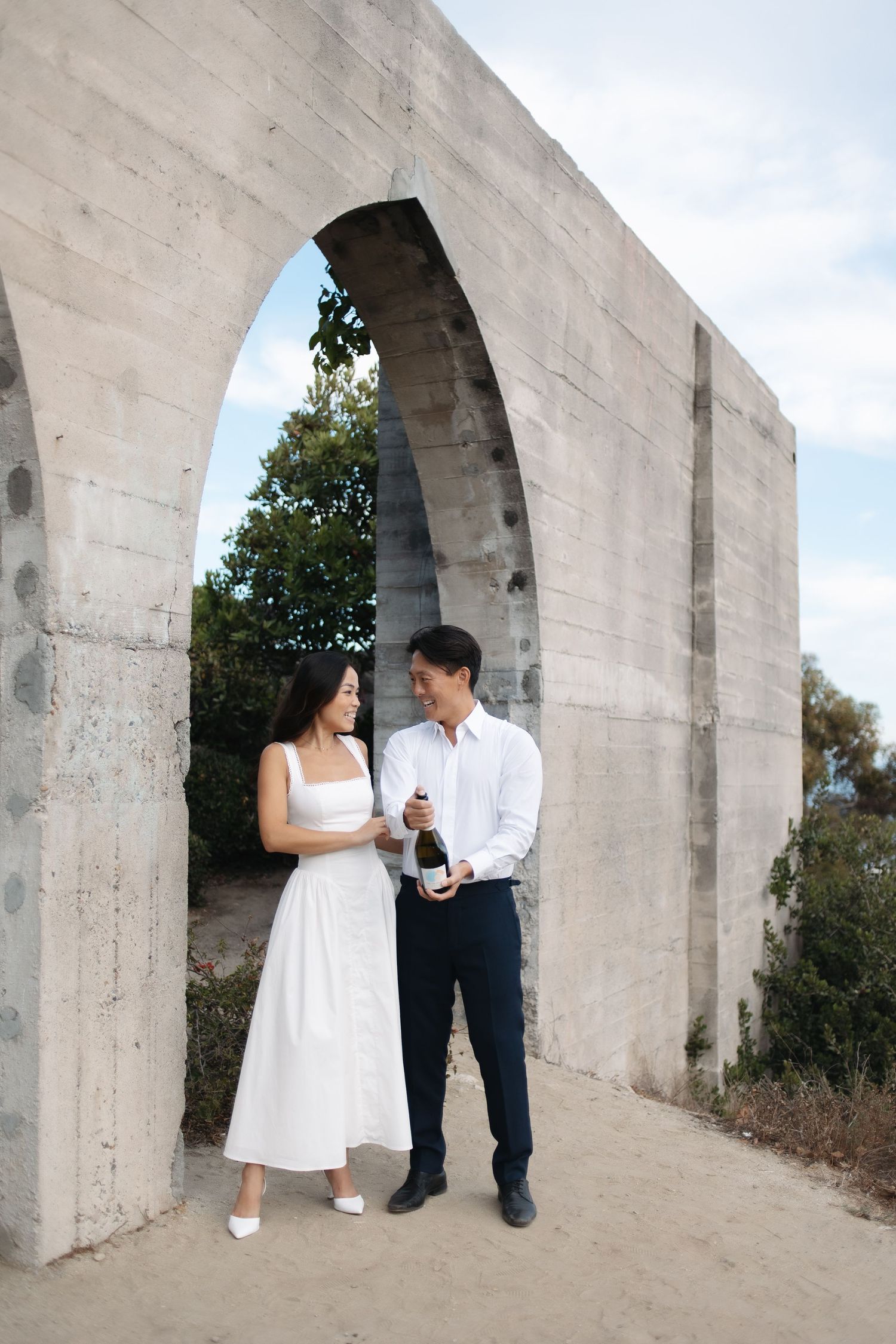 A couple holding a bottle of champagne stands under a concrete archway outdoors.