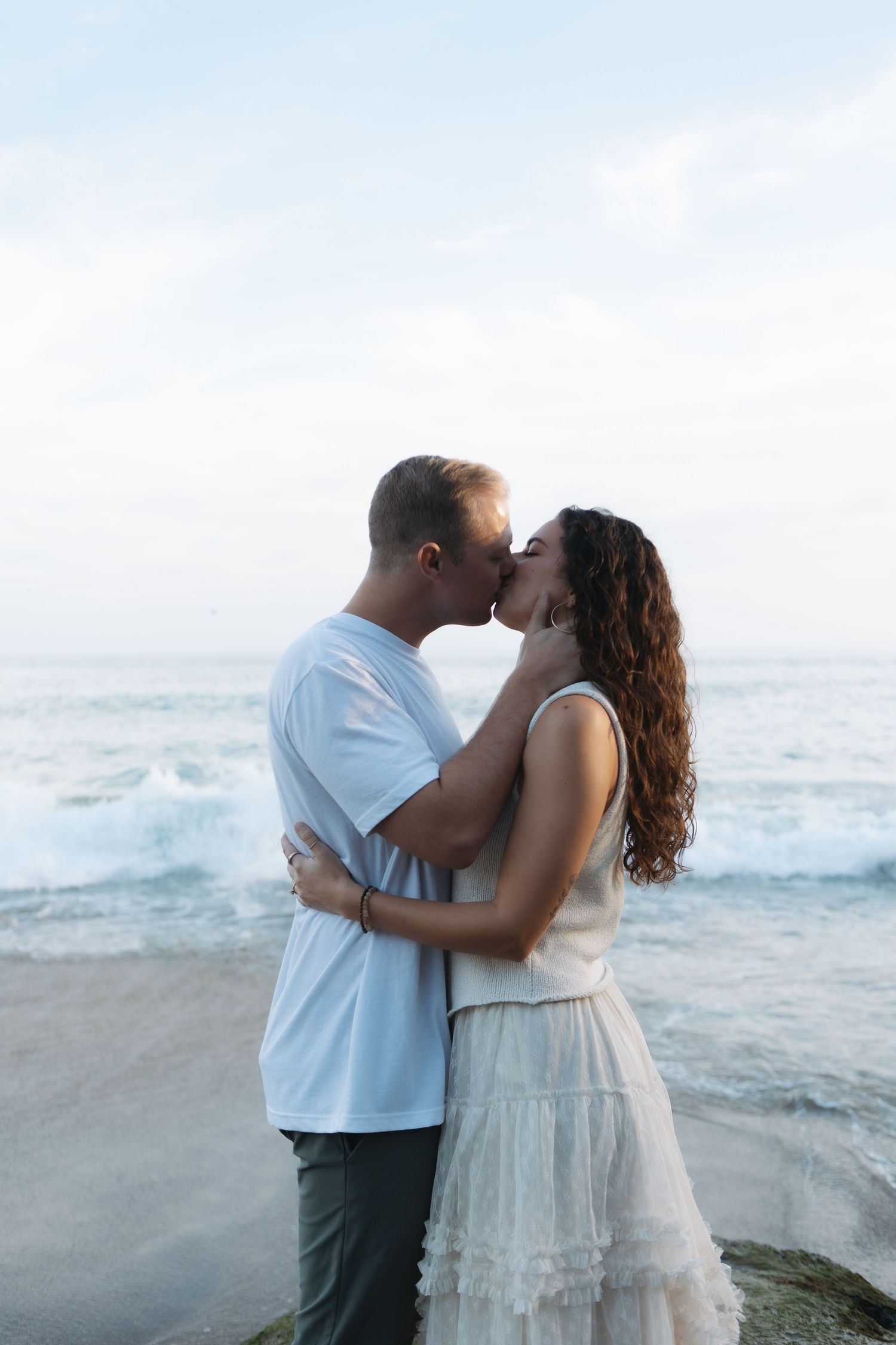 A couple shares a romantic moment on a rocky beach with crashing waves in the background.