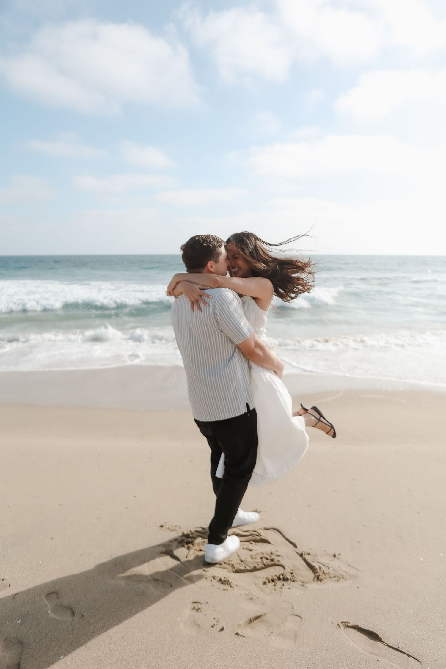 A romantic couple in white and black attire dances and spins joyfully together on a sandy beach by the ocean.