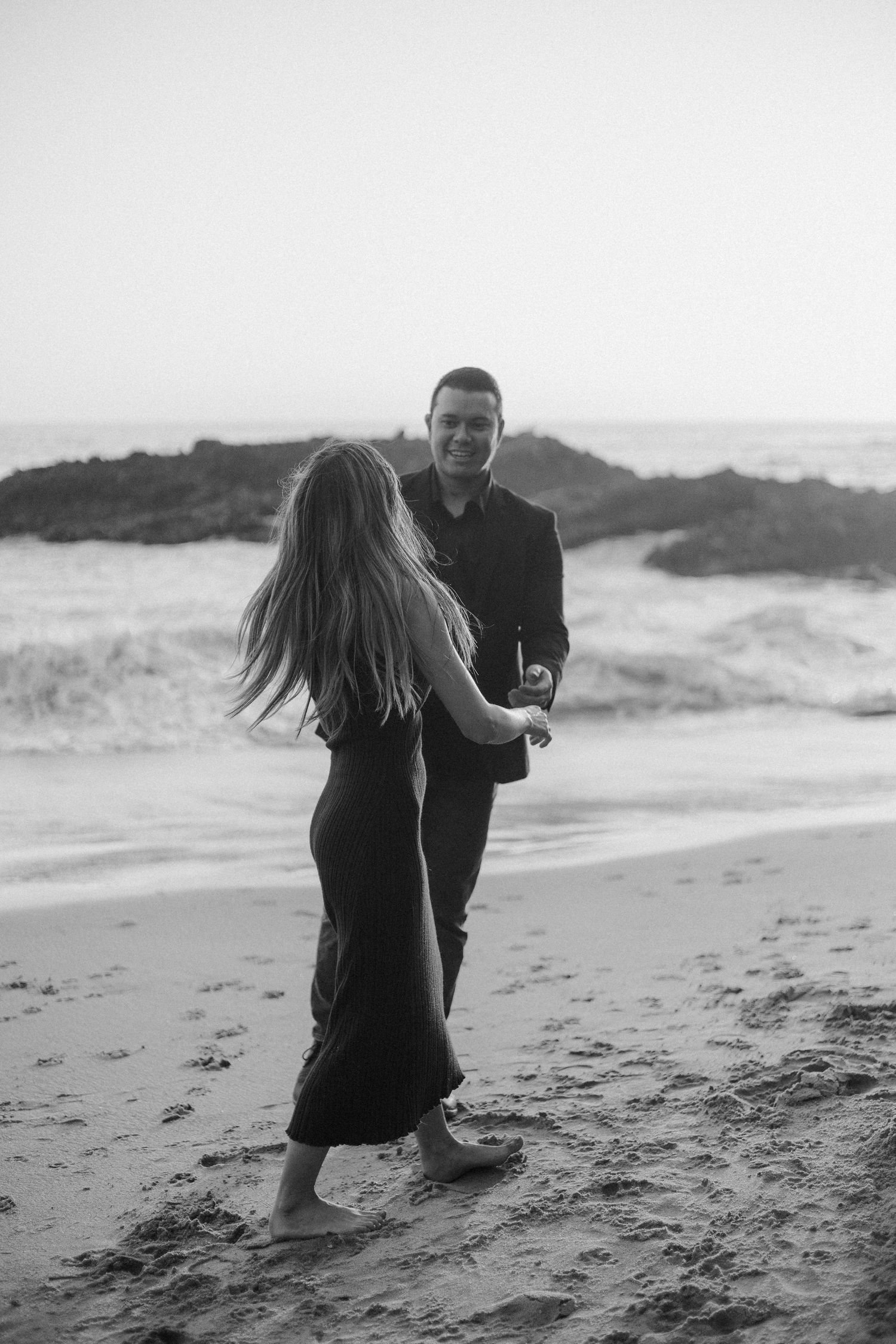 A couple in black outfits dancing together on a misty beach during sunset.