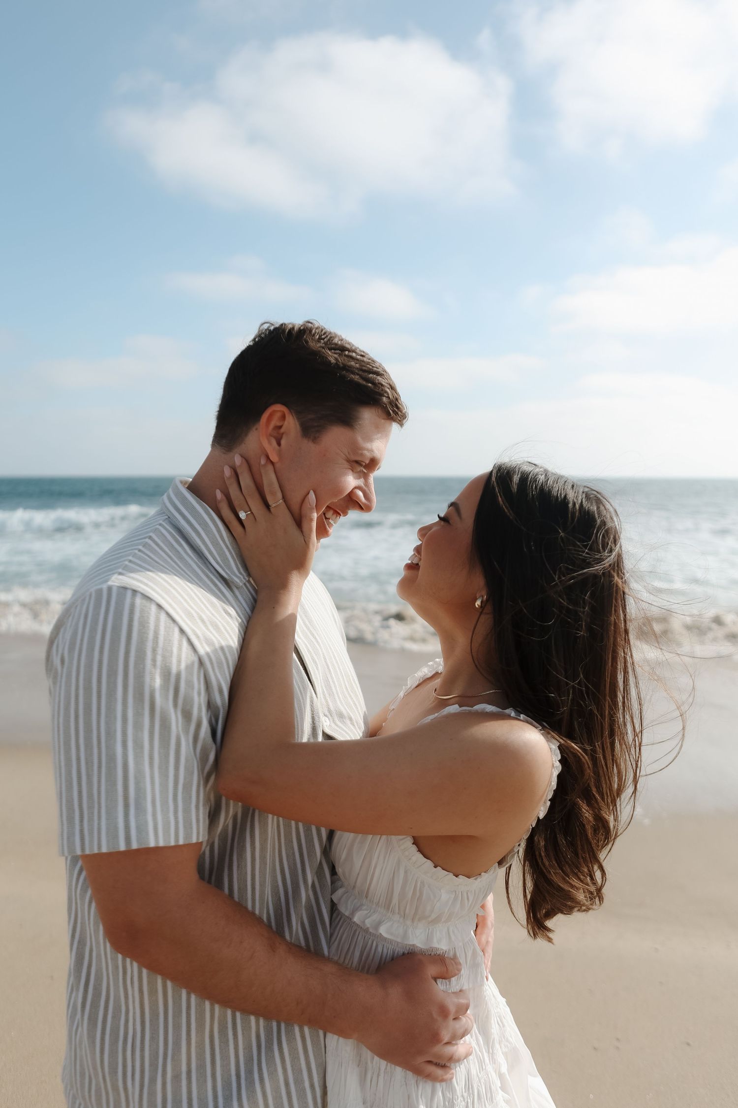 A romantic couple embraces and laughs together on a sunny beach with ocean waves in the background.