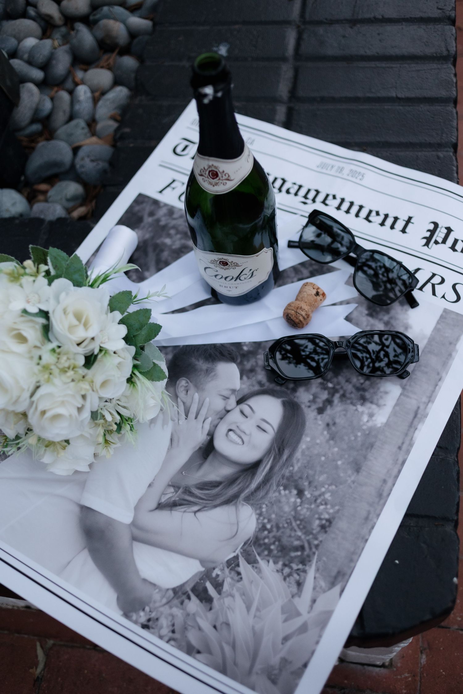 Wedding memorabilia display with champagne bottle, white roses and newspaper on dark surface.