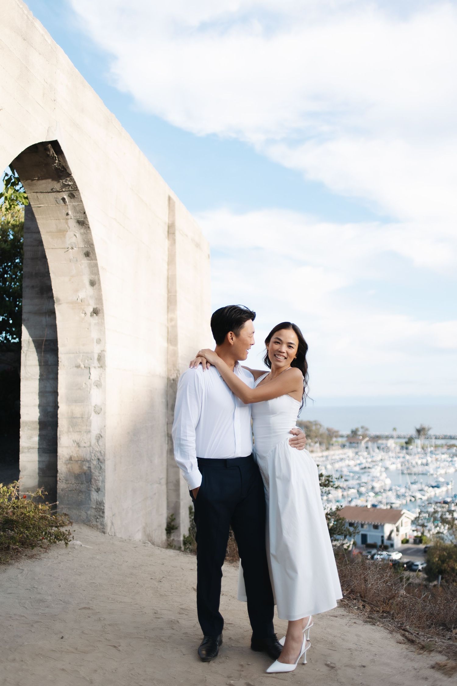A romantic couple in white attire embraces against stone archways overlooking a coastal Mediterranean cityscape.