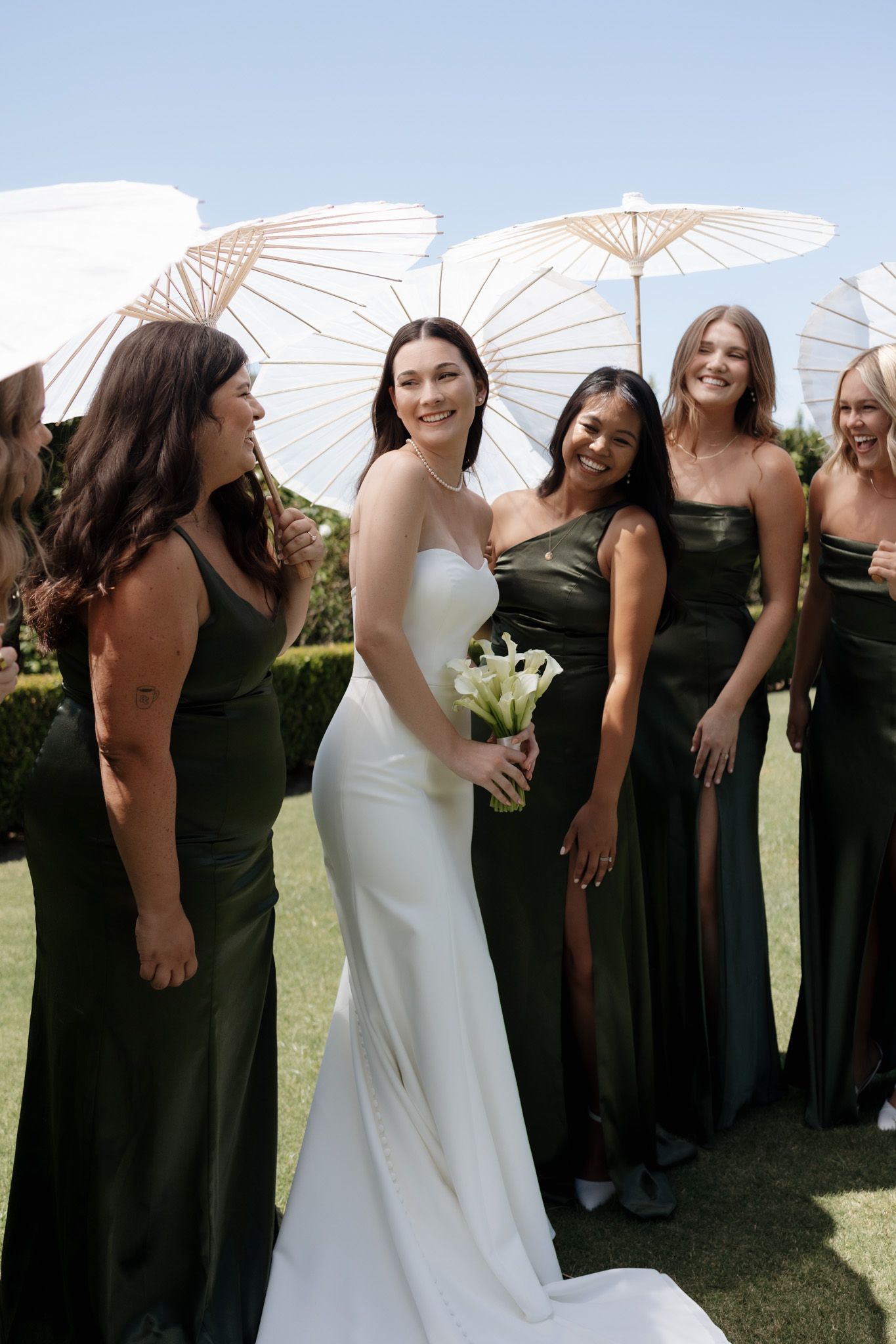 A wedding party in black bridesmaid dresses and white bridal gown pose playfully with umbrellas outdoors.