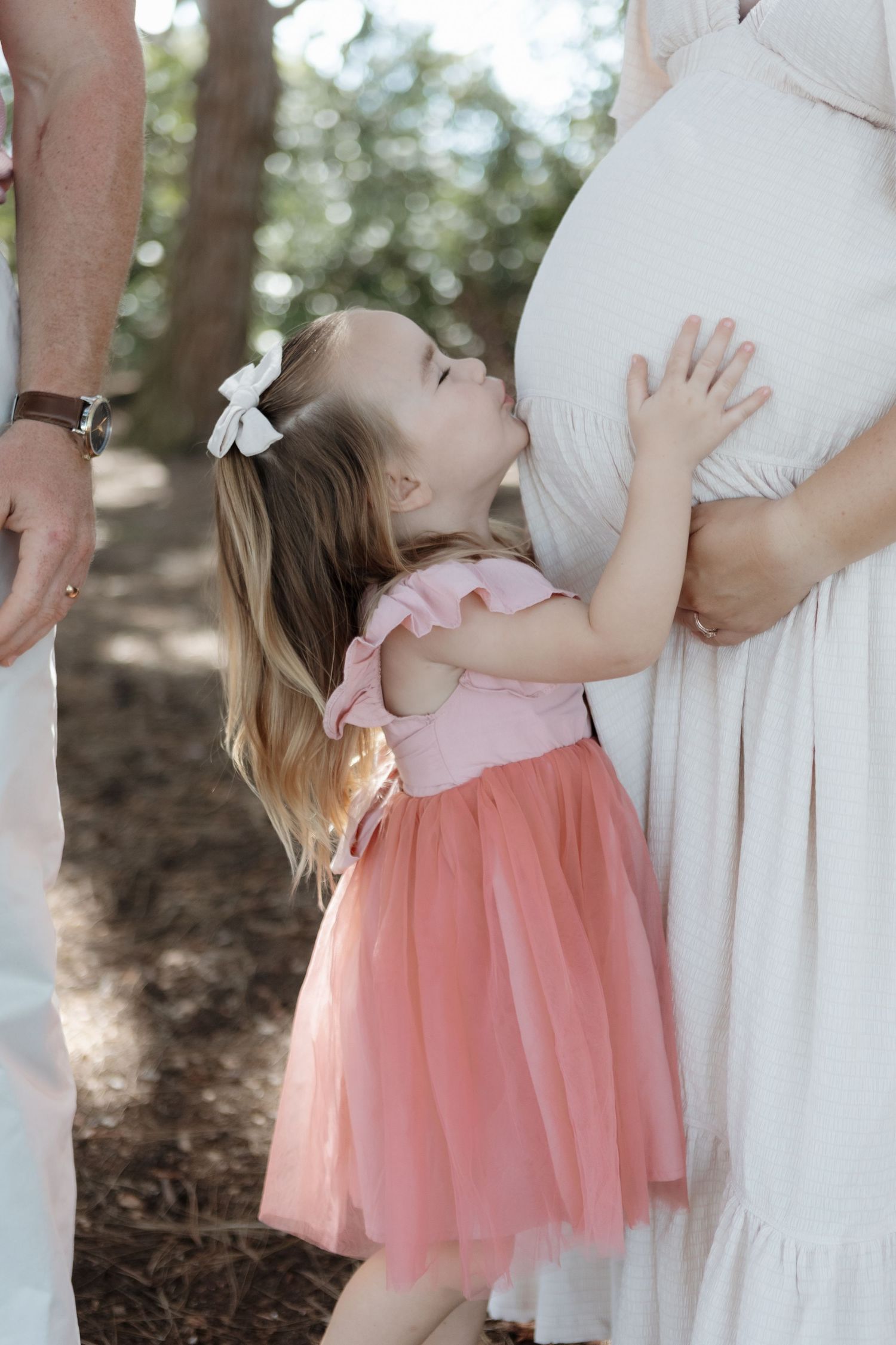 Little girl in a pink tulle dress leans back against a white dress in an outdoor setting.