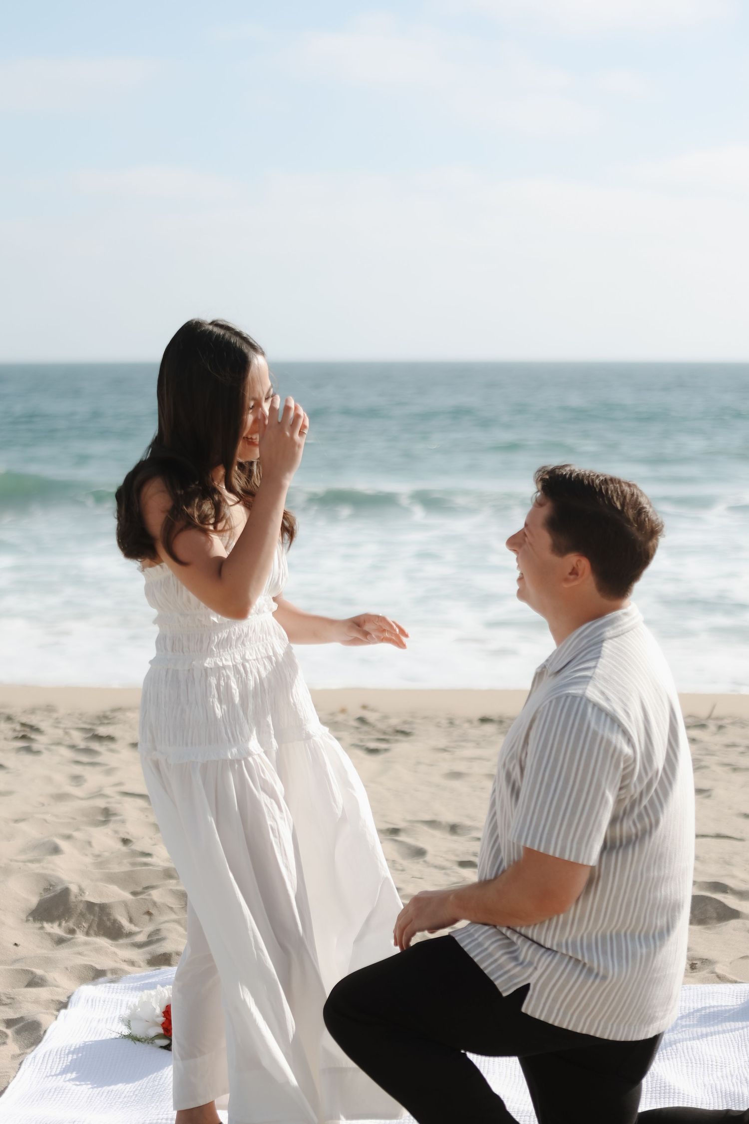 A romantic beach proposal scene with a white sundress and striped shirt against the ocean waves and sandy shore.