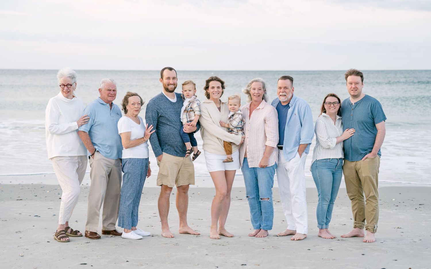 Large multigenerational family posing together on beach, smiling, dressed in coordinated light blue and white outfits.