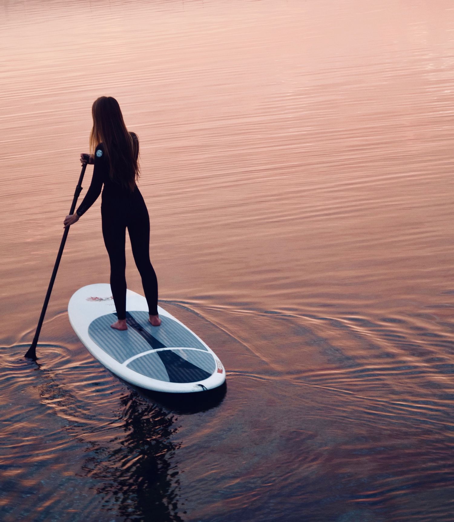 Woman in black wetsuit paddle boarding on calm pink-hued water at sunset.