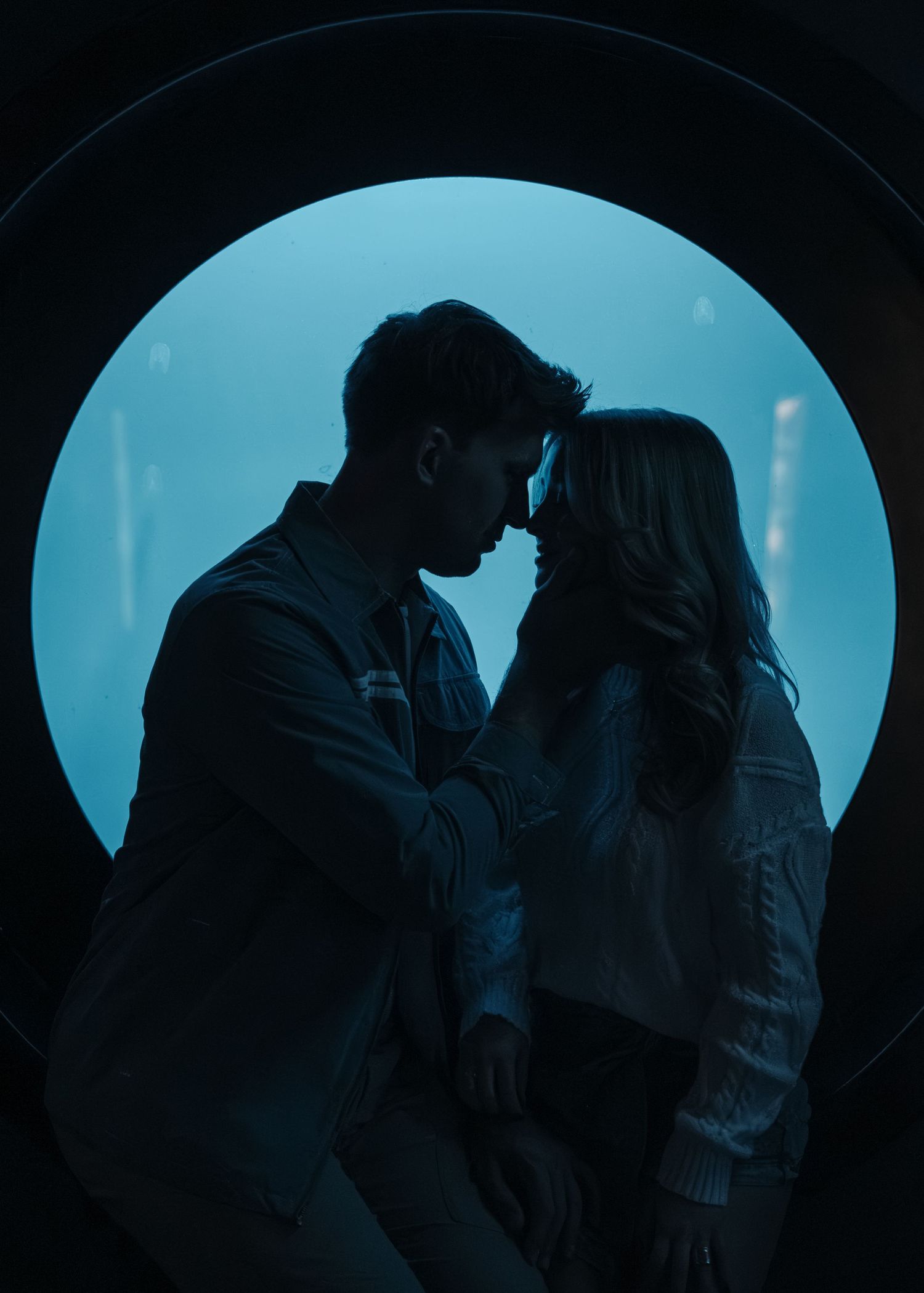 Romantic couple silhouetted in circular window sharing intimate moment in dark blue aquarium setting.