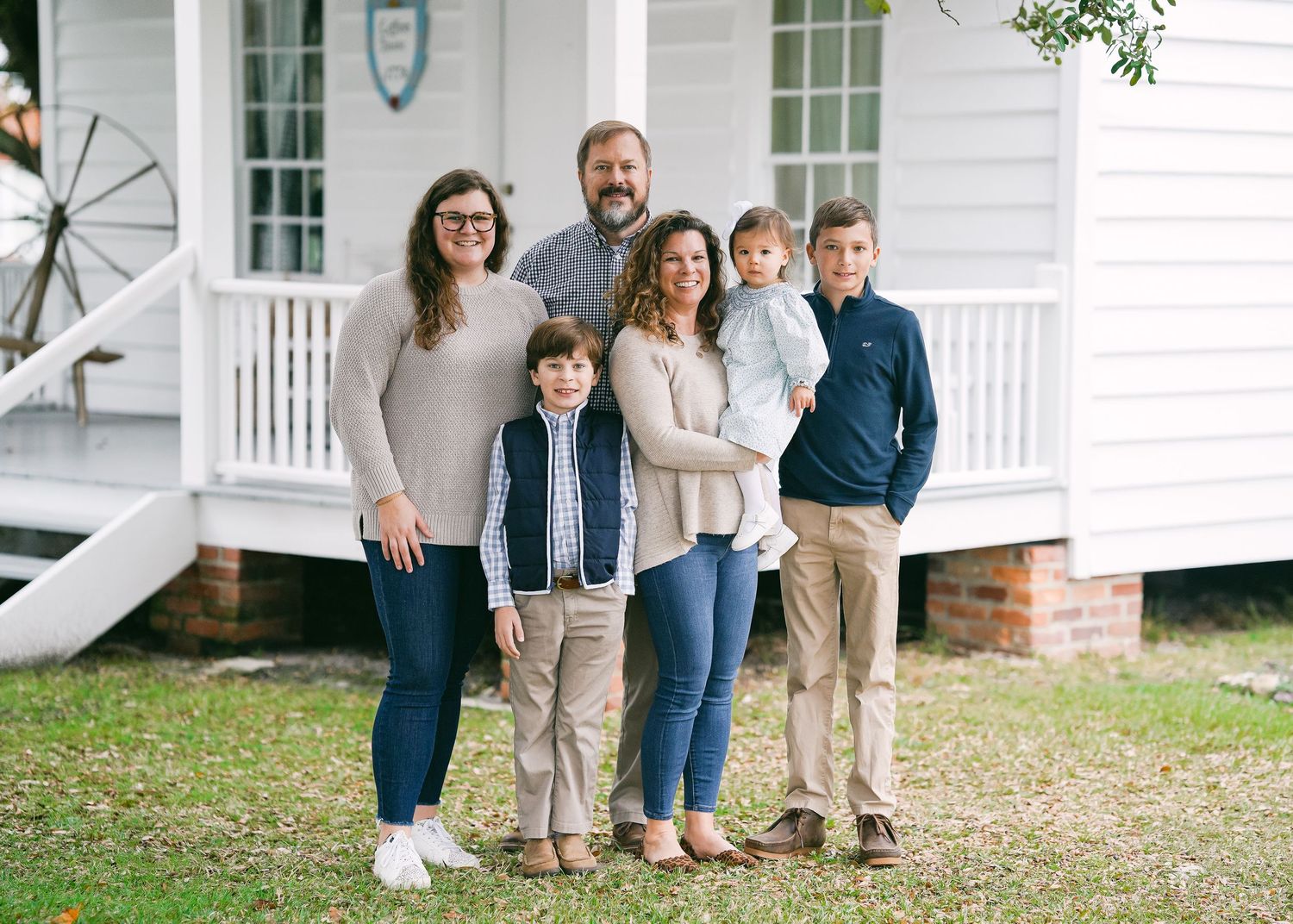 Family of five smiling in front of charming white farmhouse on sunny day.