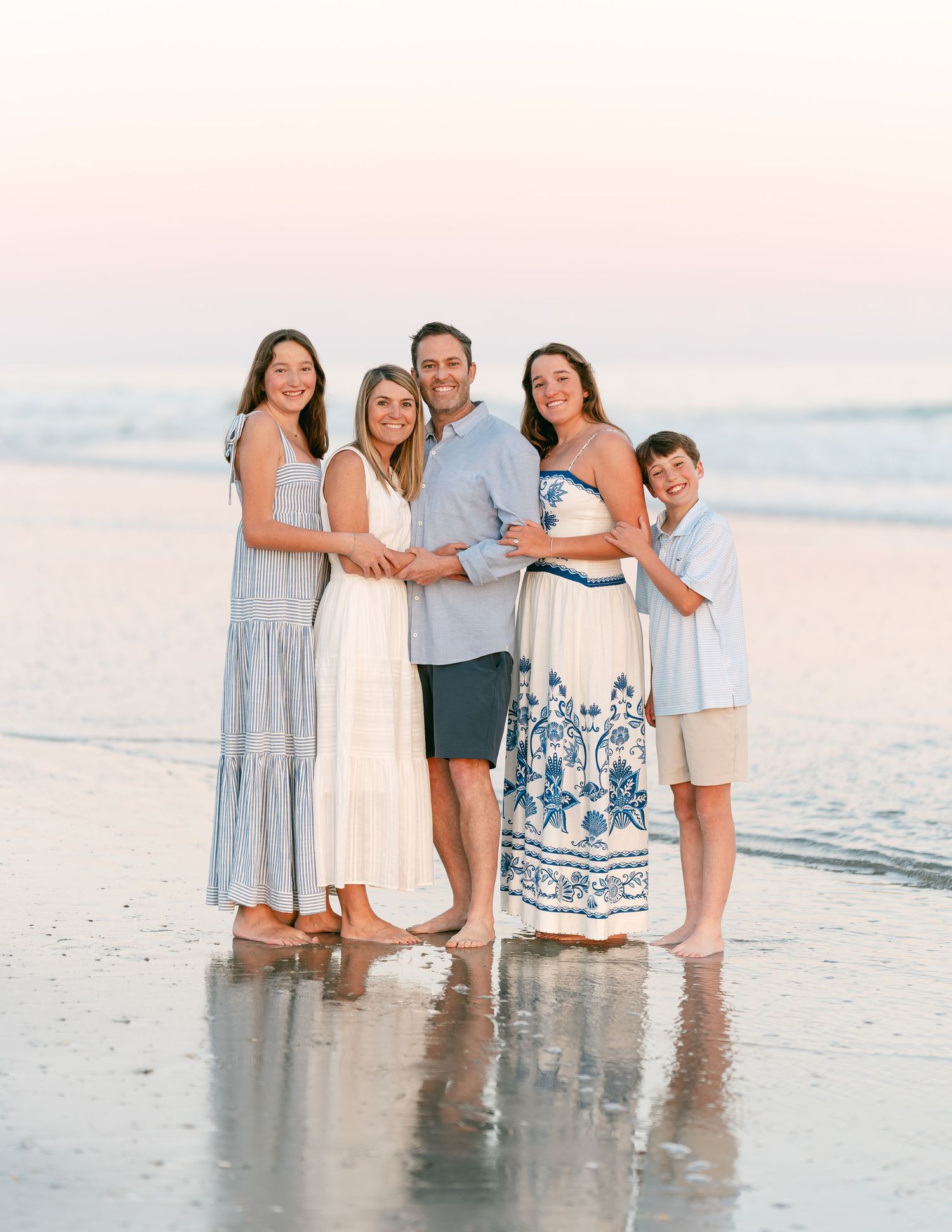 Joyful family of five posing together on reflective beach at sunset in coordinated outfits.