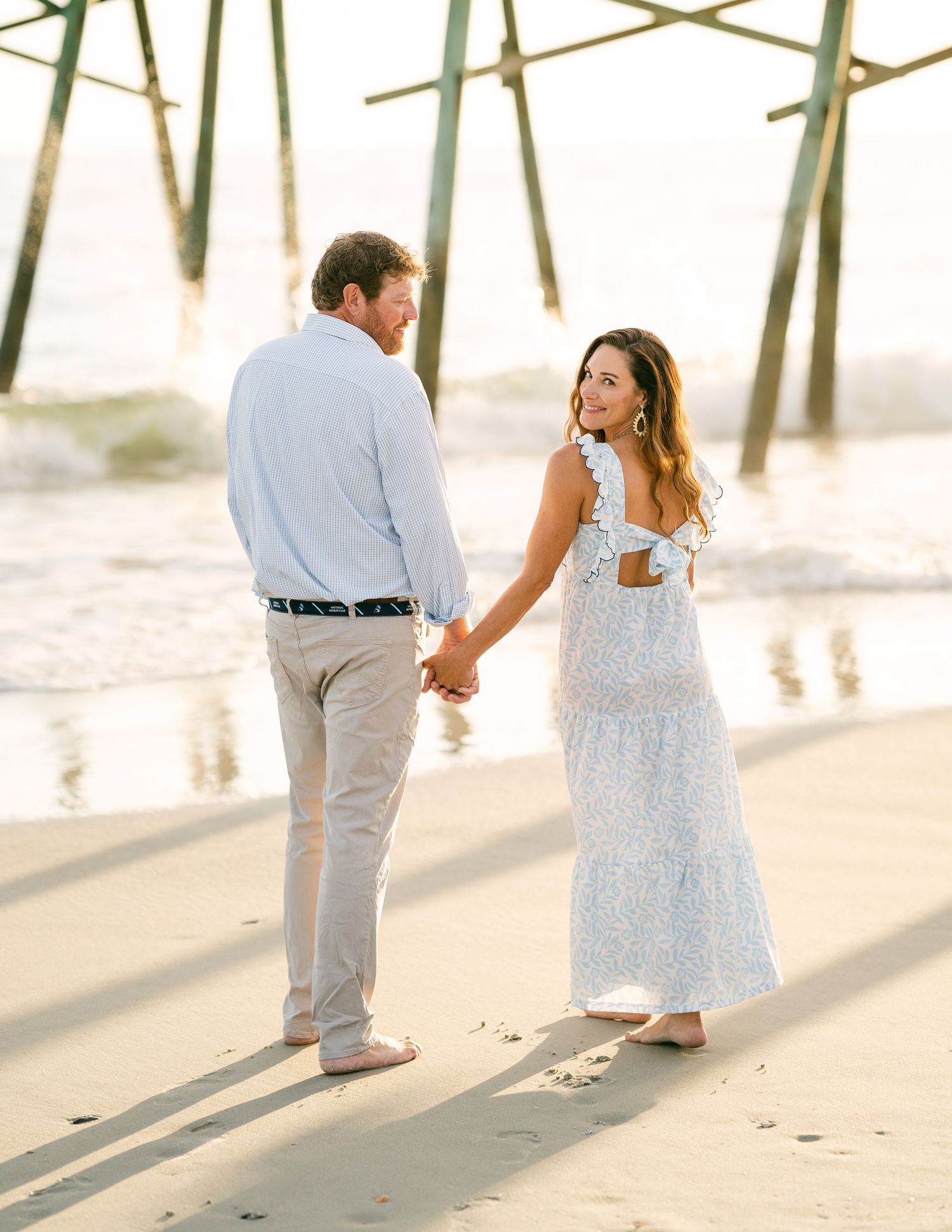 Couple holding hands on beach near pier at golden hour.