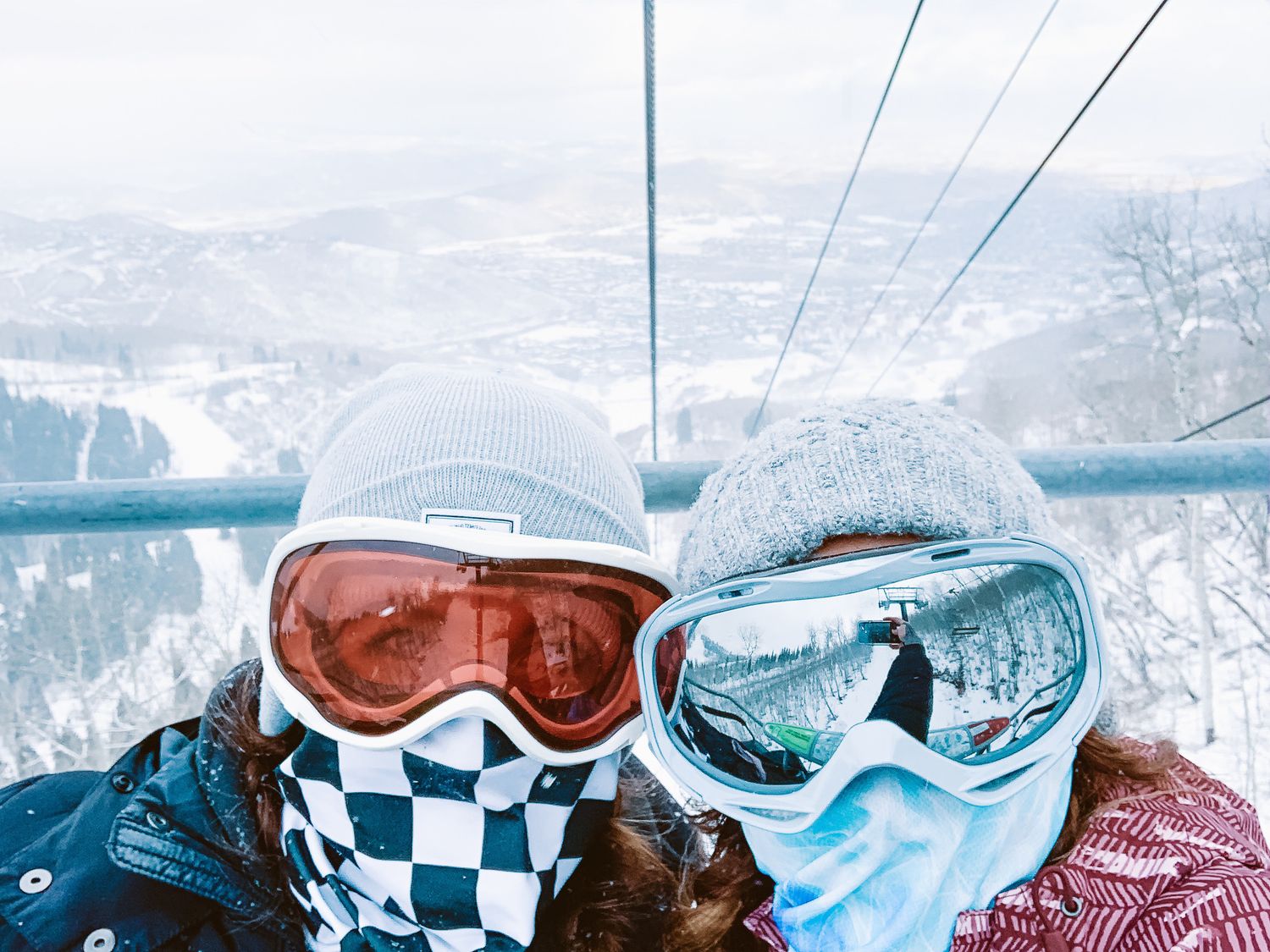 Two skiers wearing colorful goggles and masks pose for a selfie on a snowy mountain ski lift.