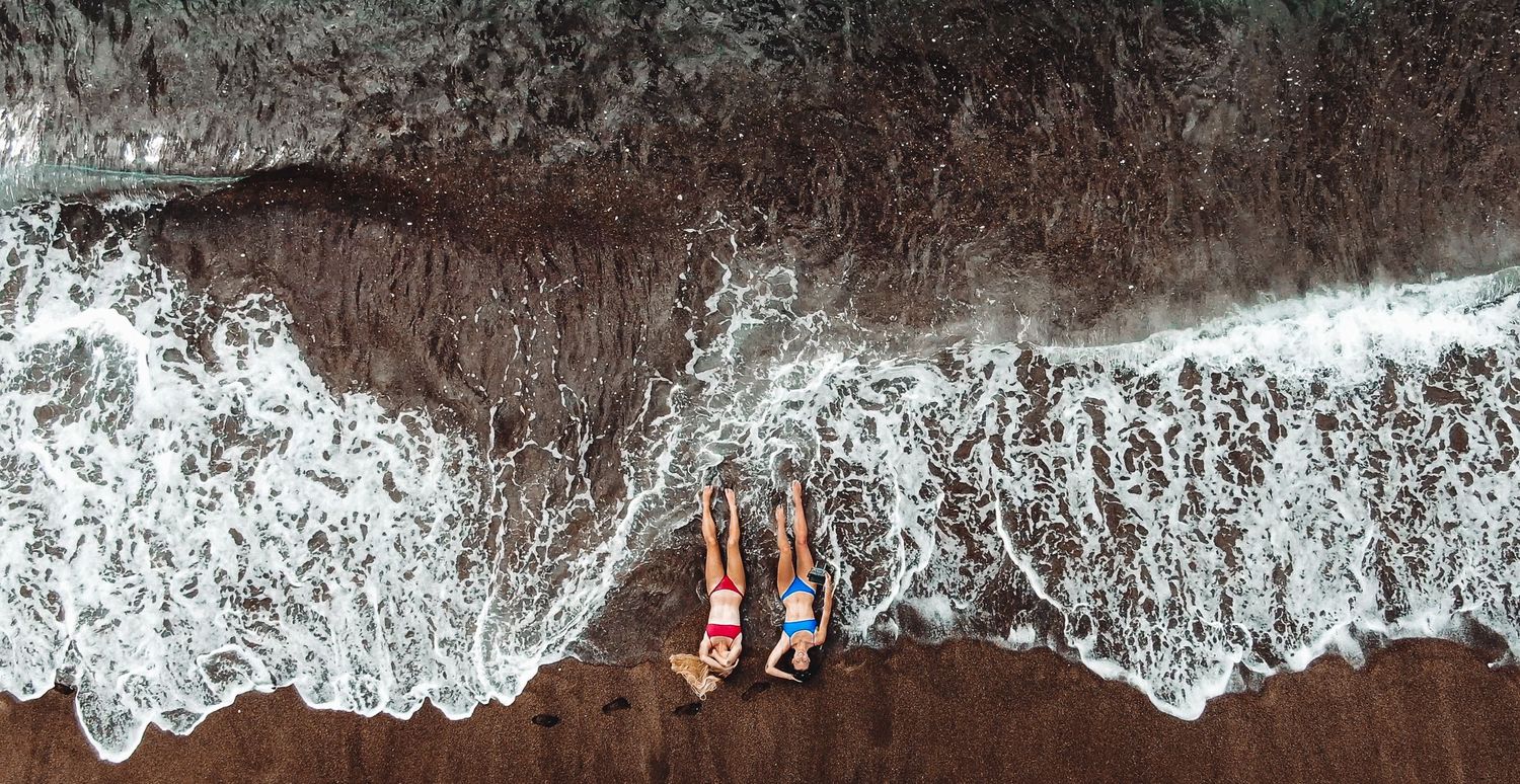 Aerial view of two people lying on dark sand beach with ocean waves.