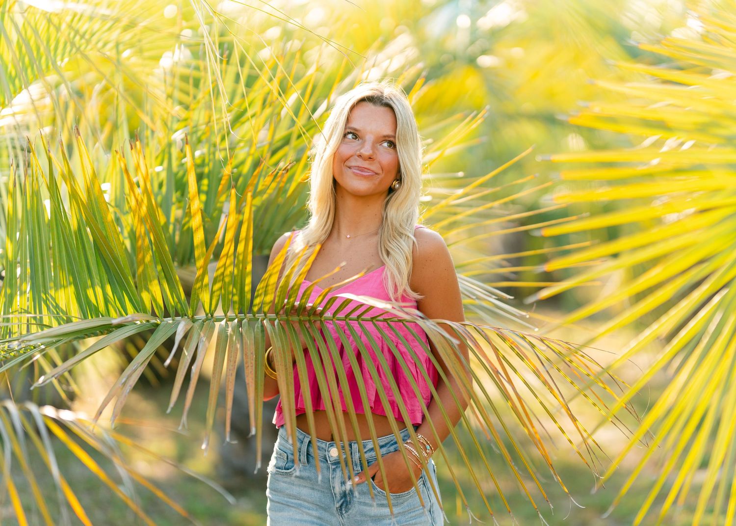 Blonde woman in pink top among tropical palm fronds in golden sunlight.