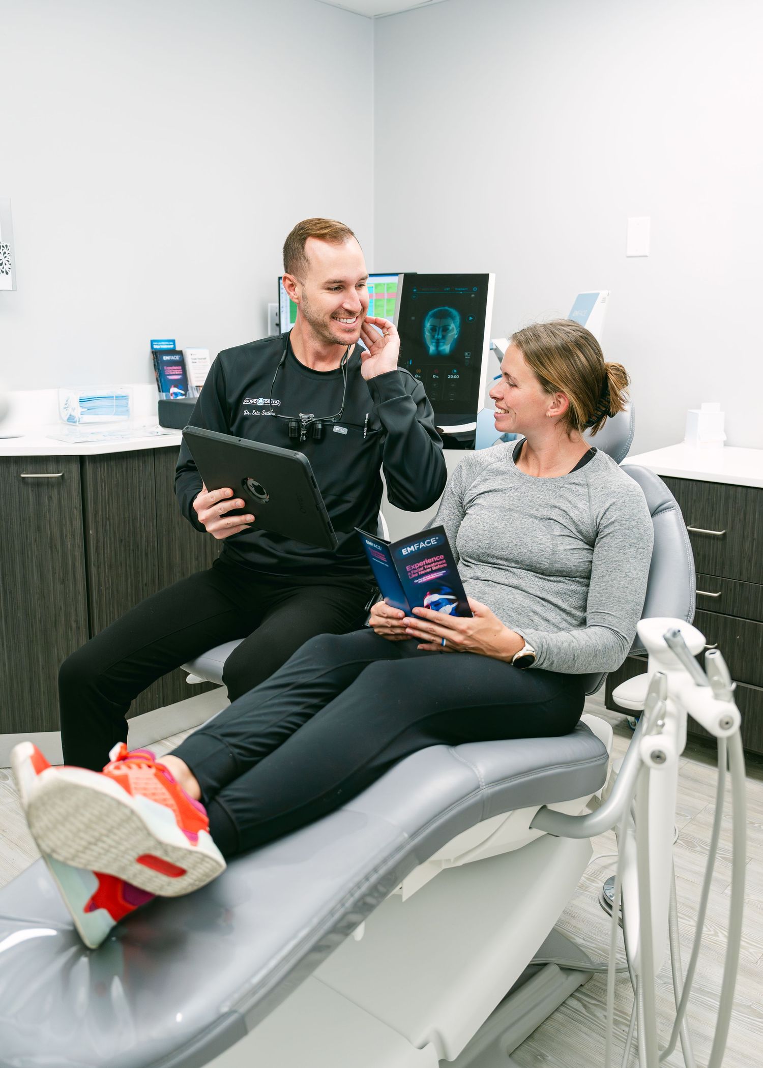 Dental professional reviews treatment plan on tablet with smiling female patient in modern dental office chair.