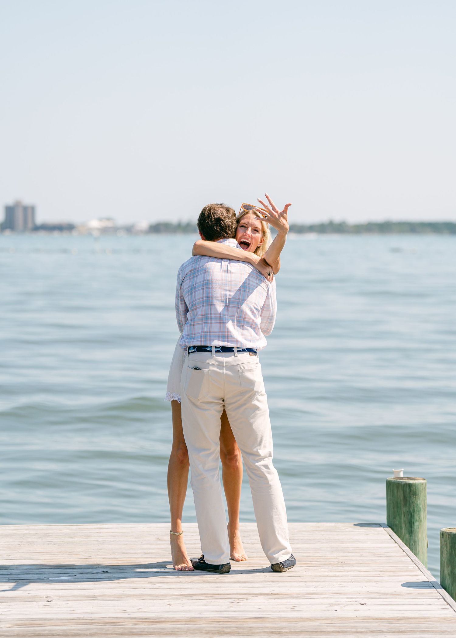 Excited woman showing off engagement ring while being lifted by partner on a sunny dock.