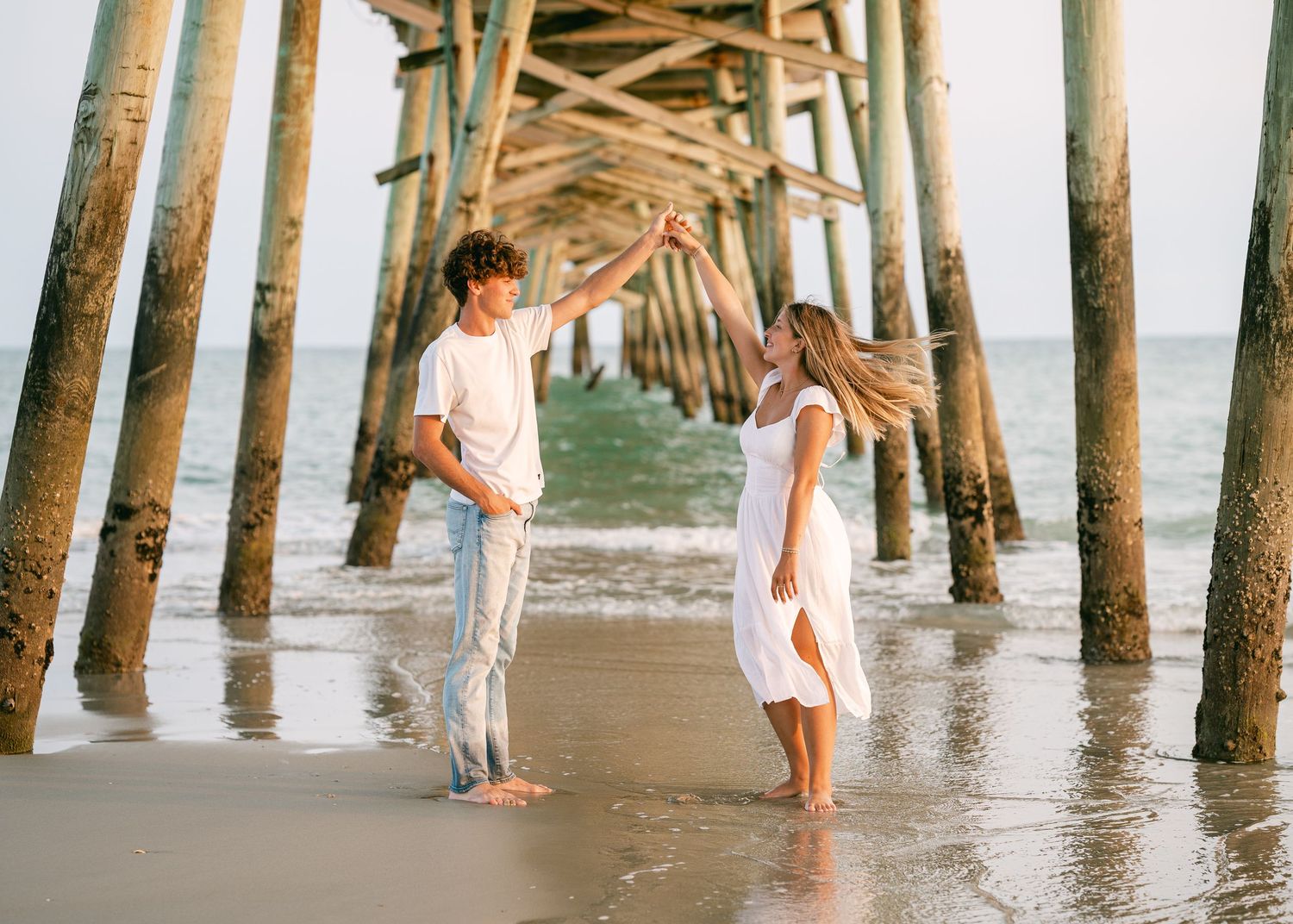 Romantic couple dancing barefoot under a wooden pier on a sandy beach at sunset.