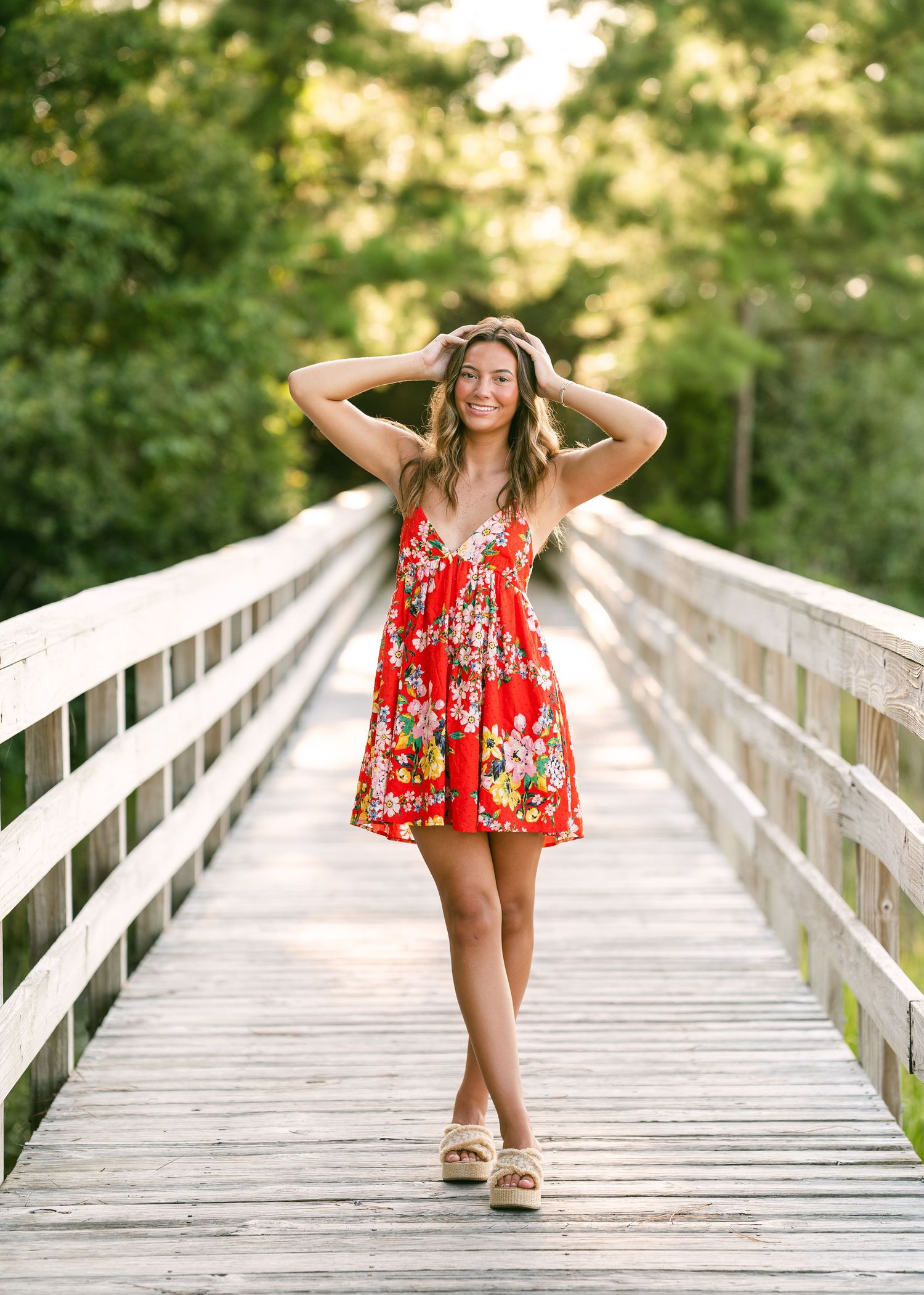 Smiling brunette woman in red floral dress posing on rustic wooden bridge at golden hour.