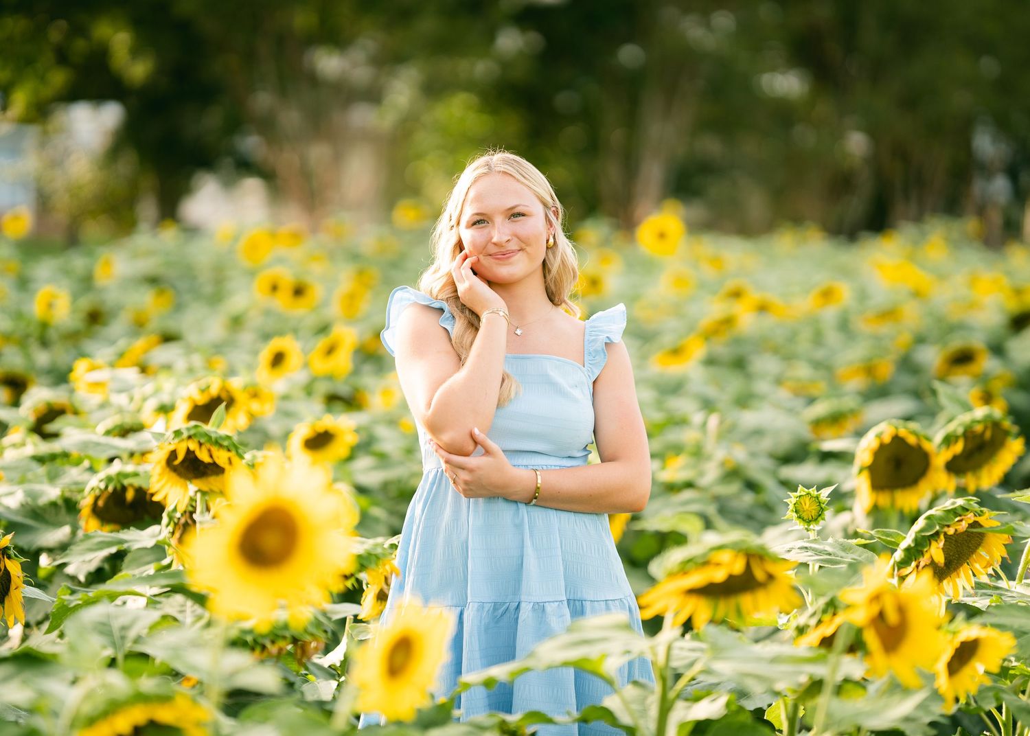 Smiling blonde woman in light blue dress posing among vibrant yellow sunflowers in a sunny field.