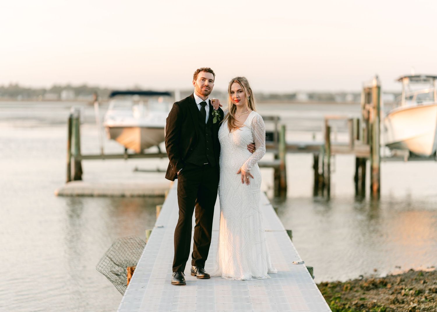 Wedding couple poses together on a dock at golden hour with boats and calm water in the background.